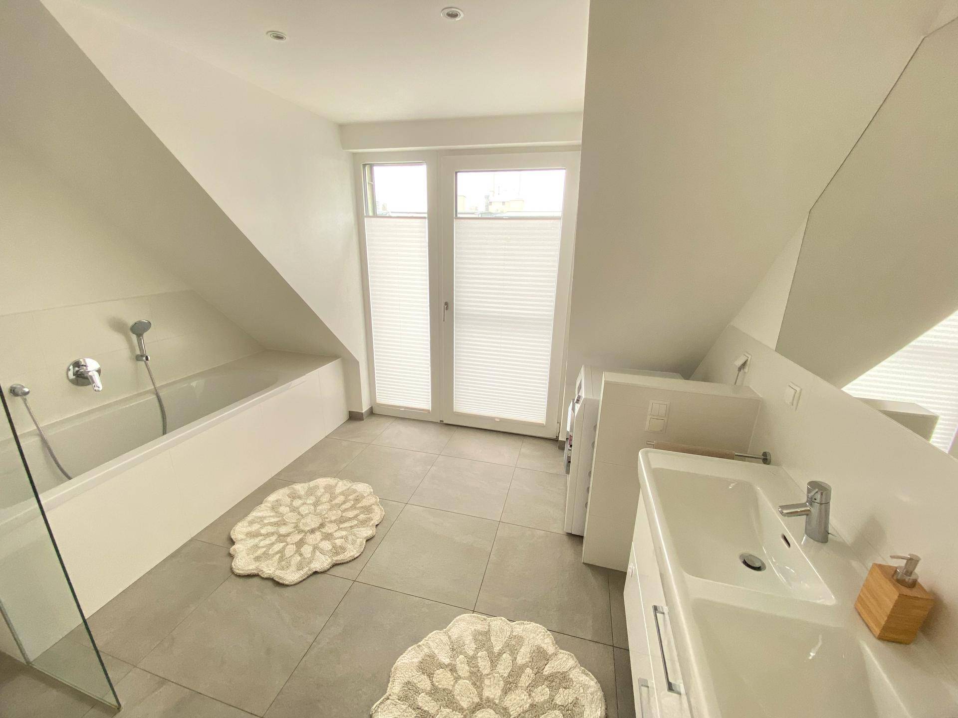 Helles Badezimmer mit Badewanne, Waschbecken, Fenster und Fliesenboden.