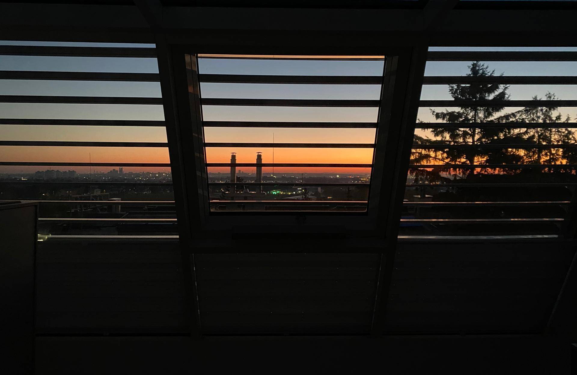 Blick aus einem Fenster auf einen farbenprächtigen Sonnenuntergang über der Stadt mit Industrieanlagen und Bäumen.