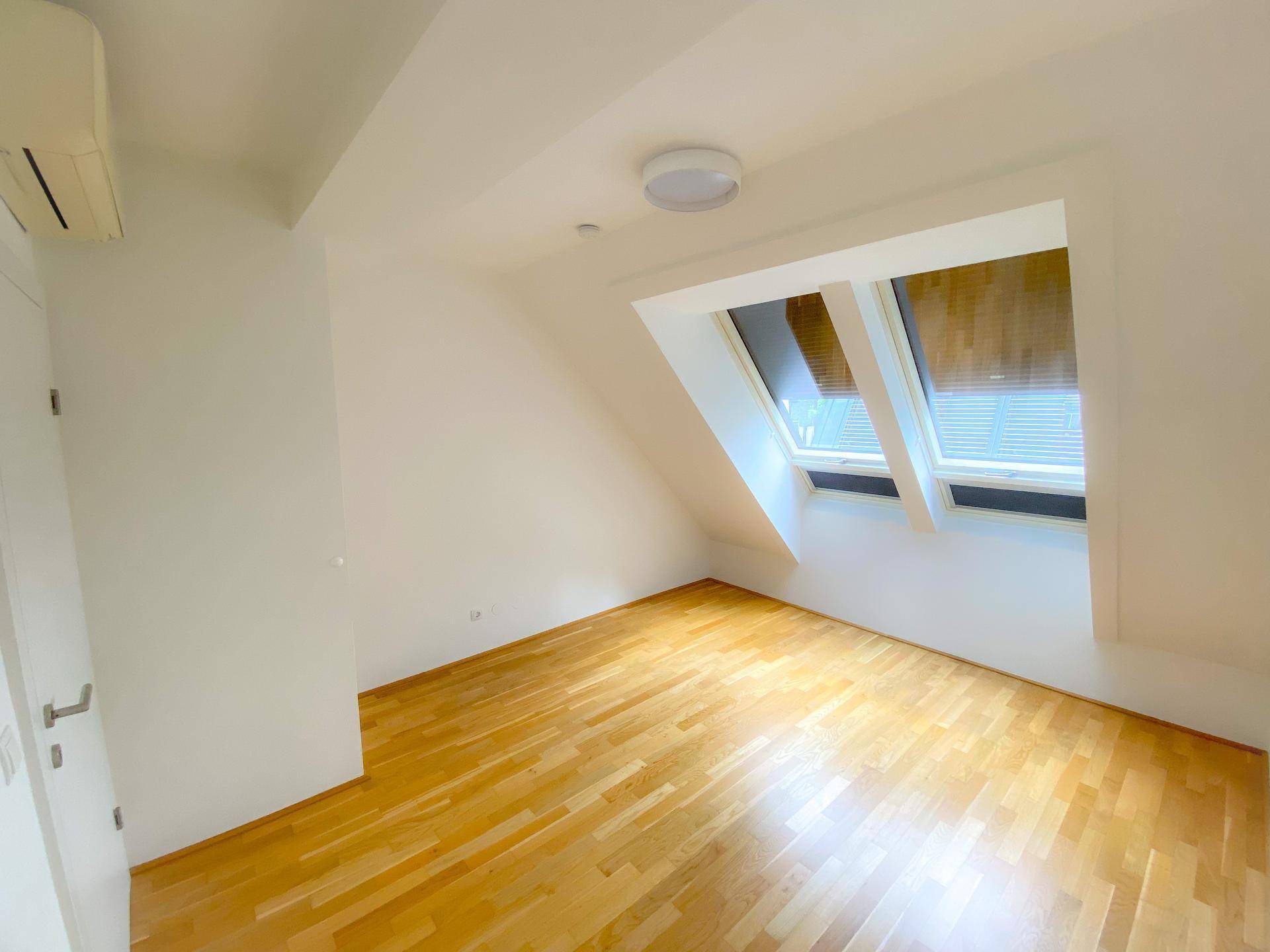 Lichtdurchflutetes Zimmer mit Holzboden, Dachschräge, Dachfenstern und Klimaanlage.