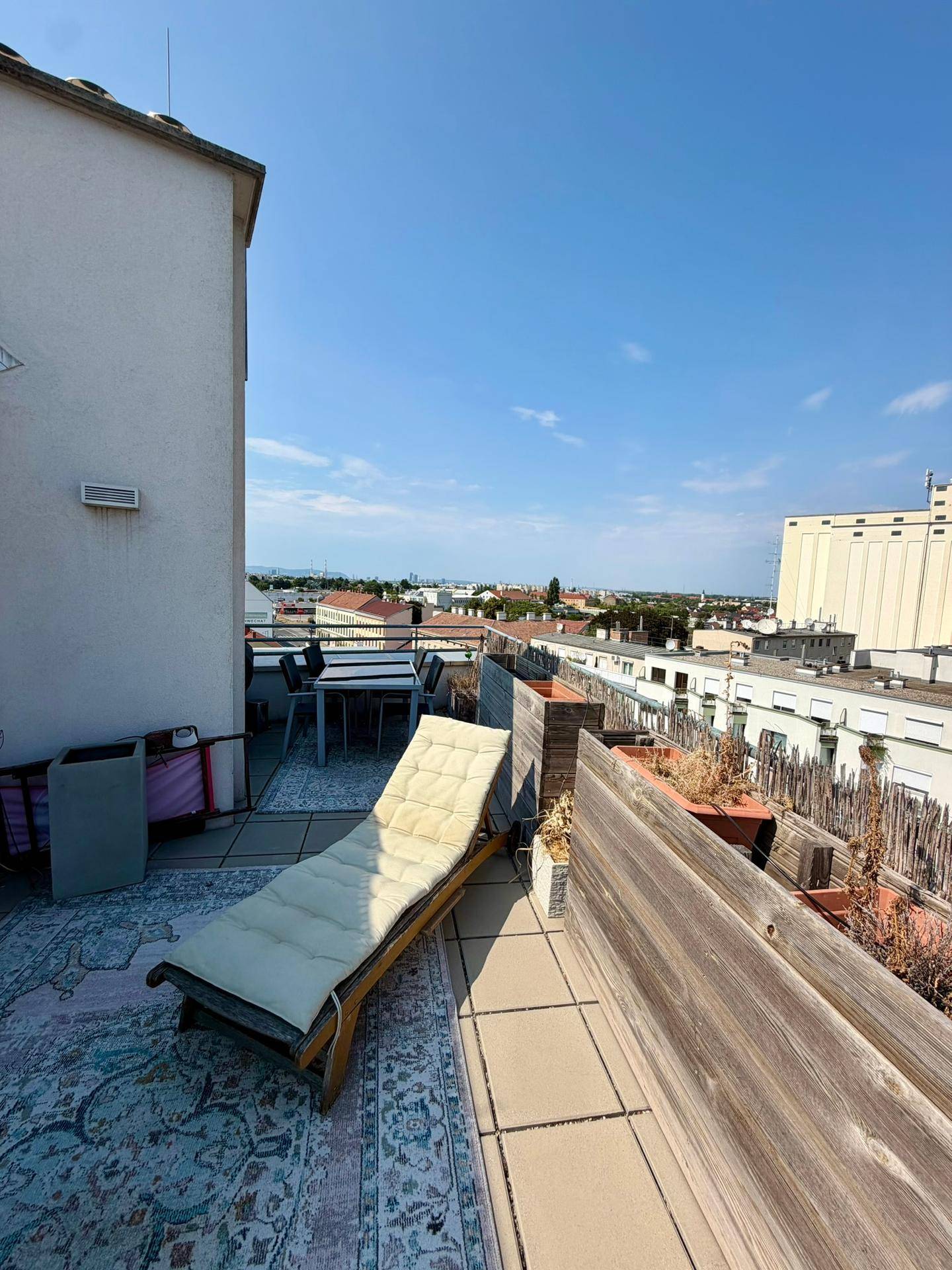 Sonniger Außenbereich auf einer Terrasse mit Liegestuhl, Esstisch und Blick über die Stadt.