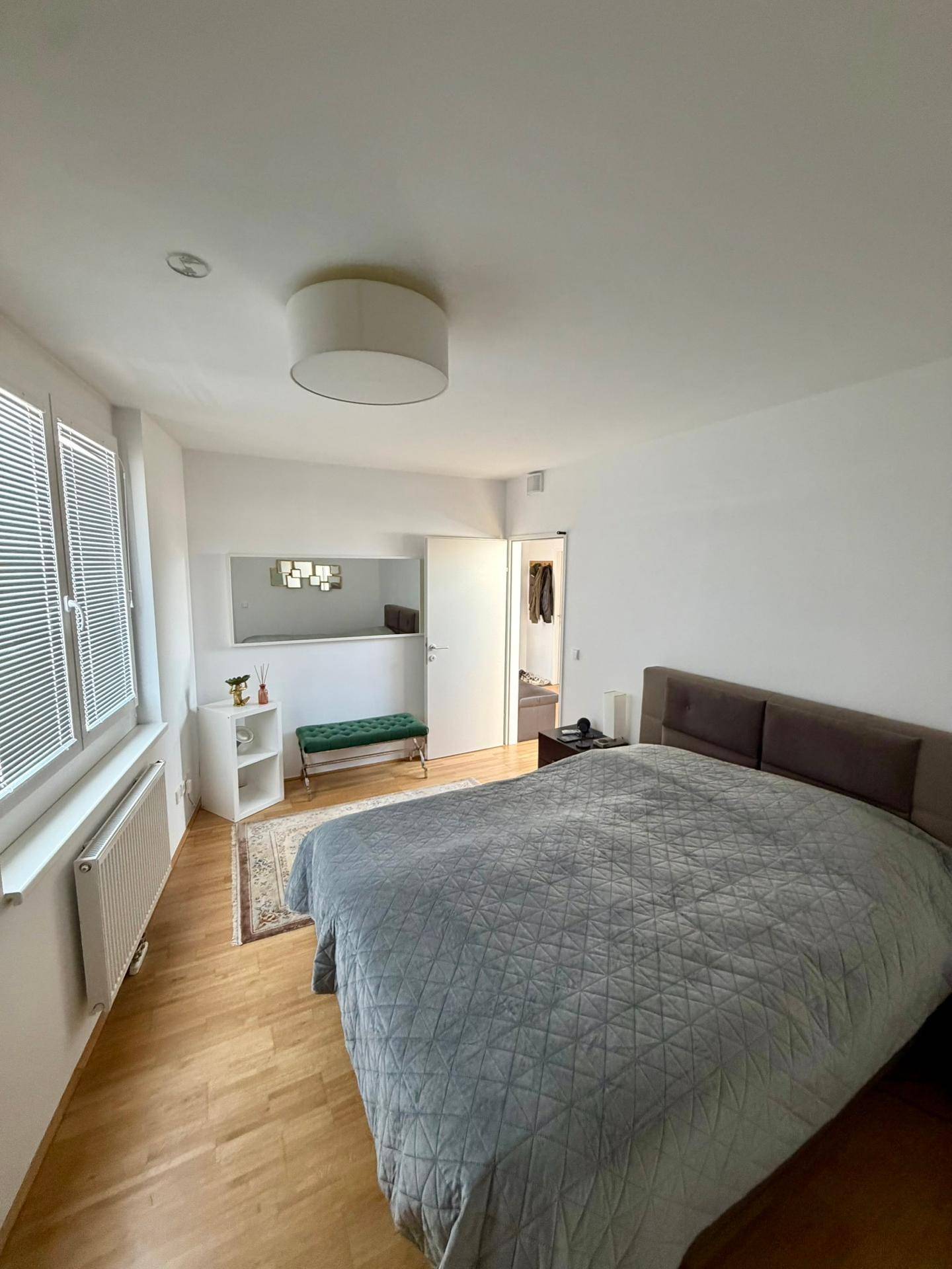 Helles Schlafzimmer mit großem Bett, Holzparkettboden und Fenstern mit Jalousien.