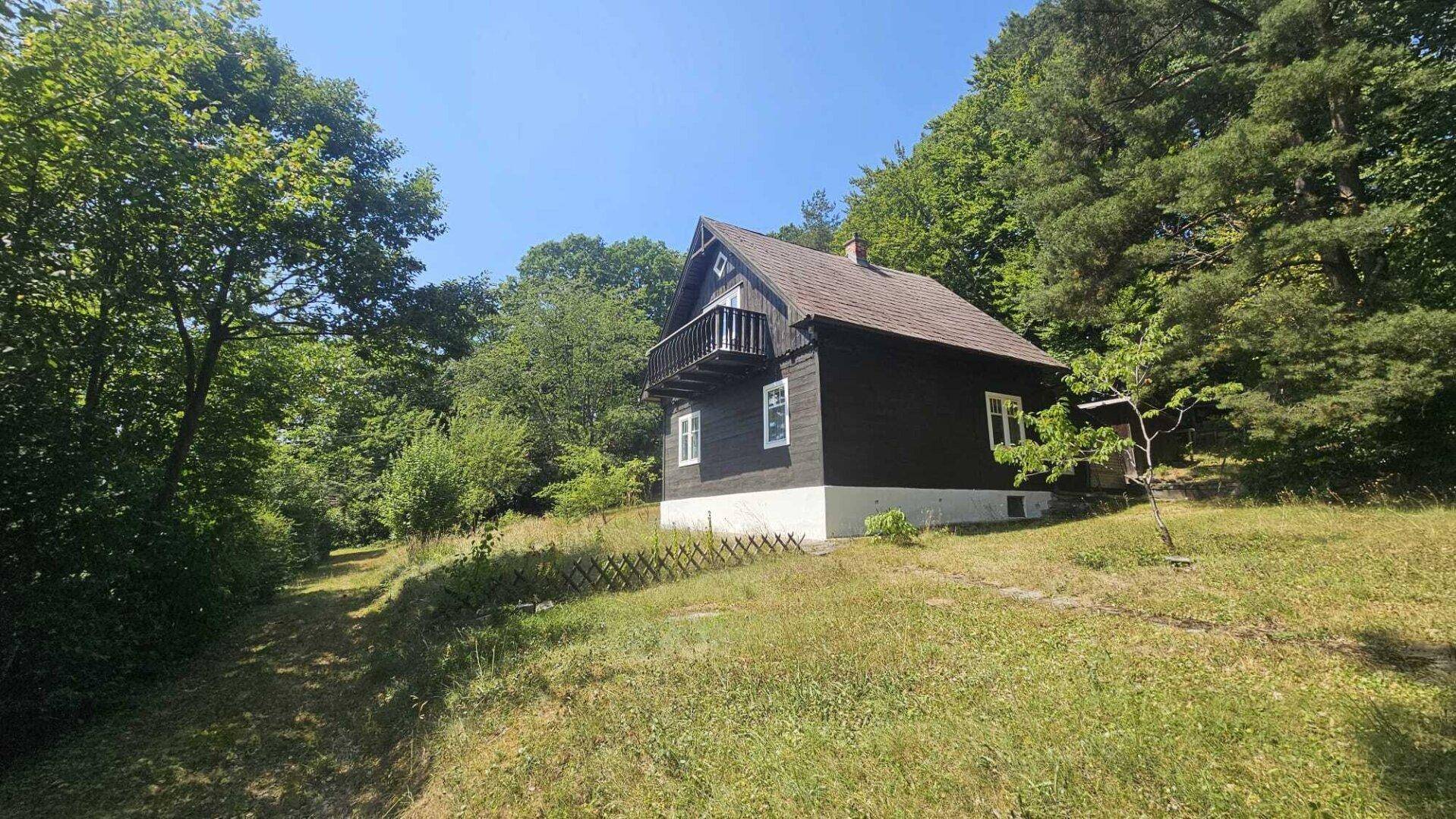 Das charmante Holzhaus mit kleinem Balkon, eingebettet in eine grüne Landschaft mit hohen Bäumen.