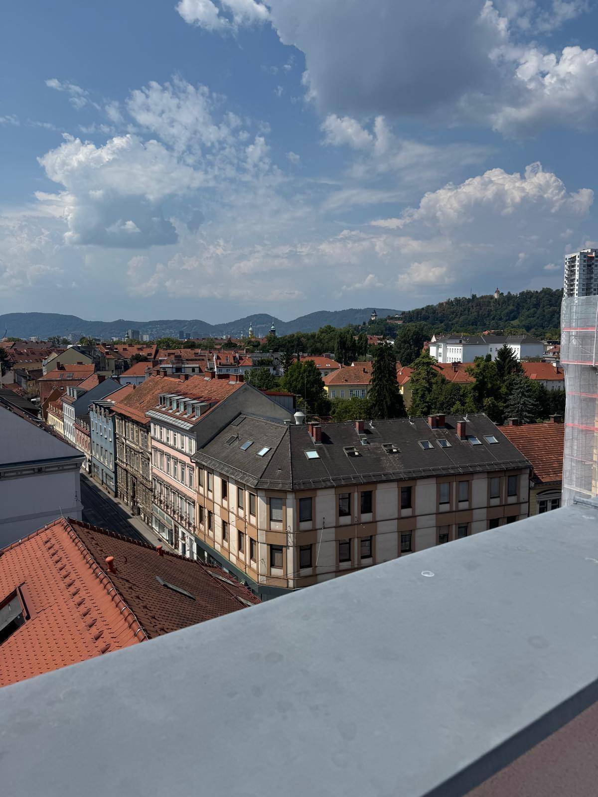 Weitläufiger Ausblick über die Stadtlandschaft mit Dächern und grünen Hügeln im Hintergrund unter blauem Himmel.