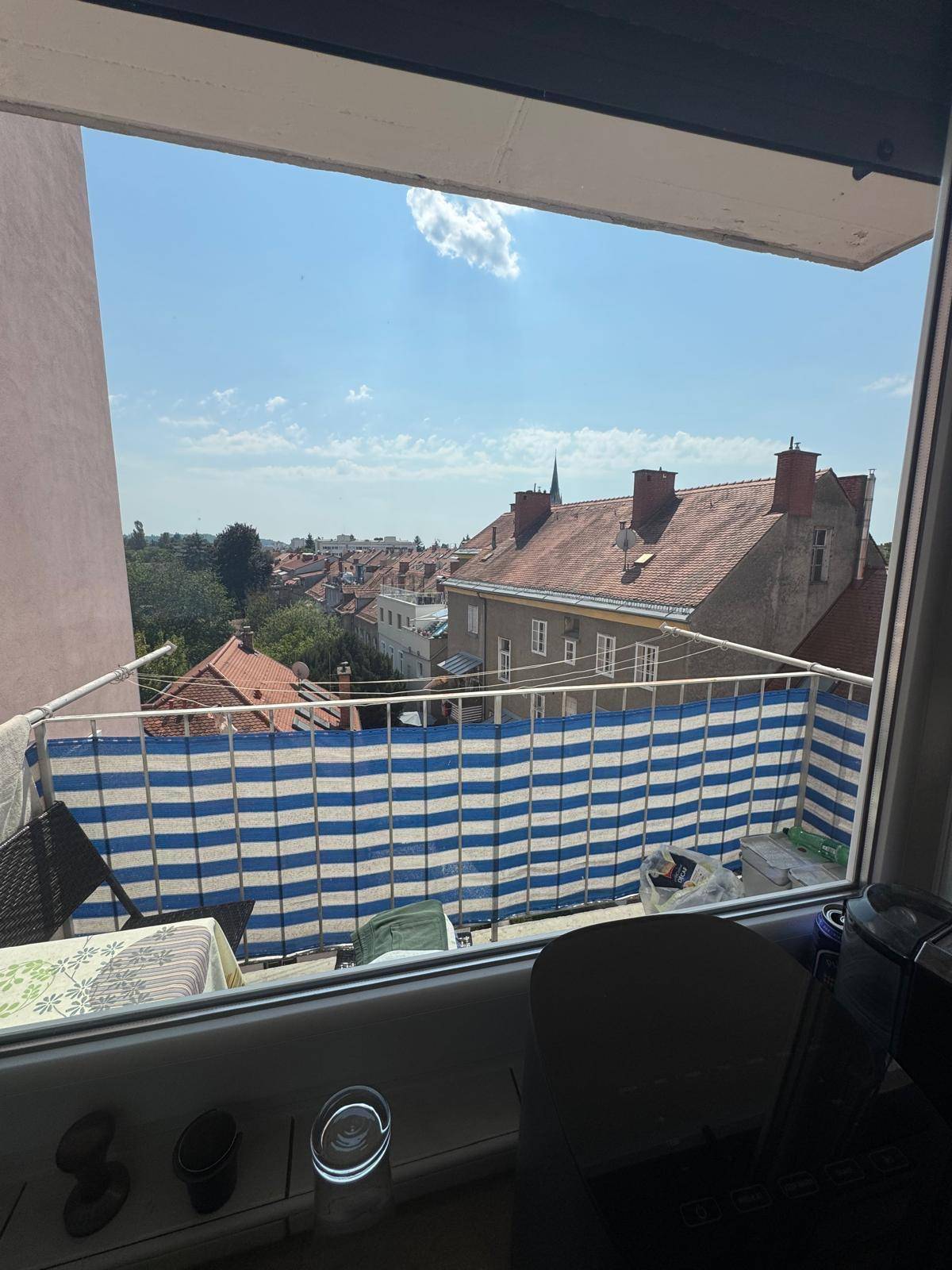 Blick vom Fenster auf den Balkon mit blau-weißem Sichtschutz und die umliegenden Dächer der Stadt.