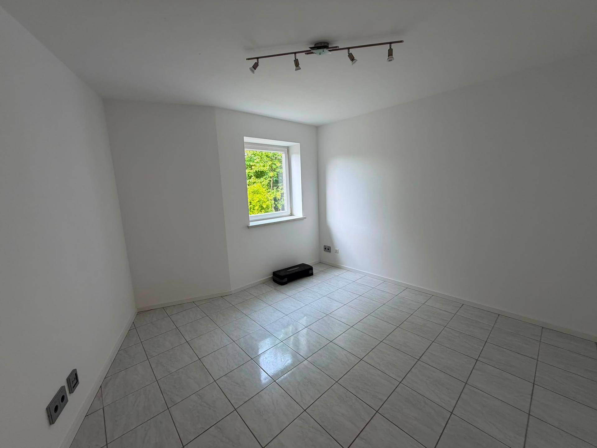 Leeres Zimmer mit Fliesenboden und Fenster mit Blick ins Grüne.