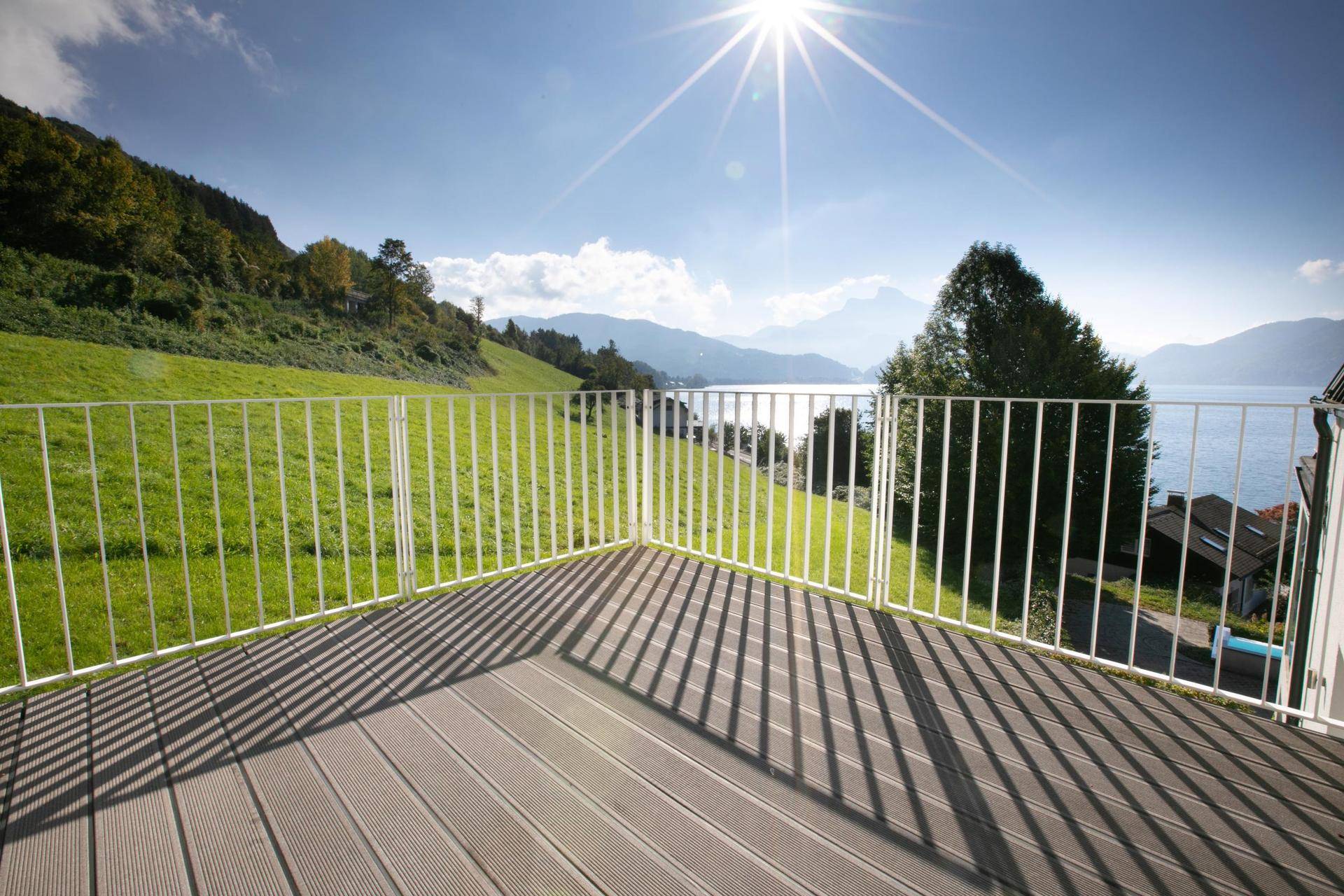 Heller Balkon mit weißem Geländer, der einen sonnigen Blick auf grüne Hügel, den See und die Berge bietet.