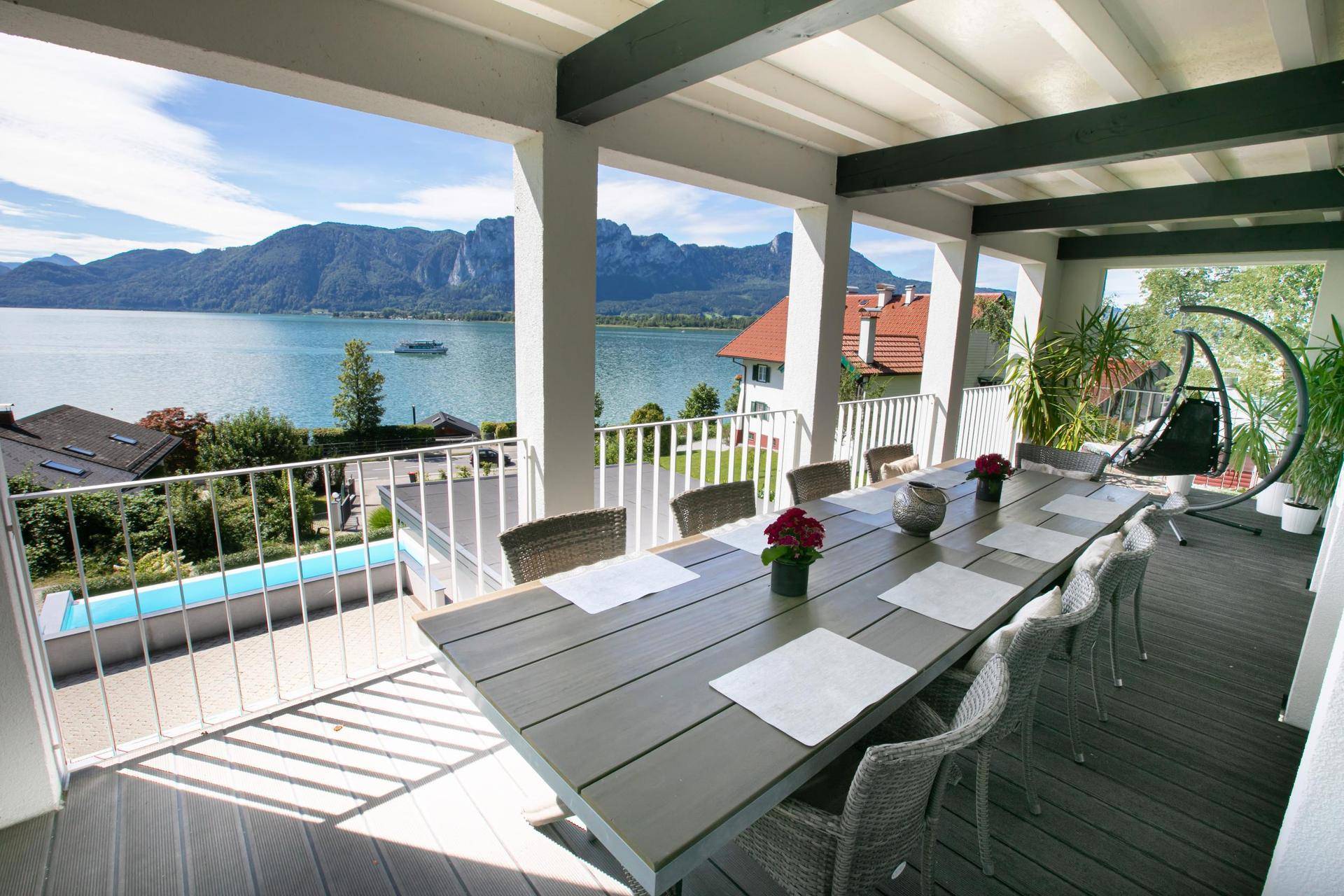 Überdachte Terrasse mit großem Esstisch und Stühlen, ideal für Mahlzeiten mit Blick auf Pool, See und Berge.