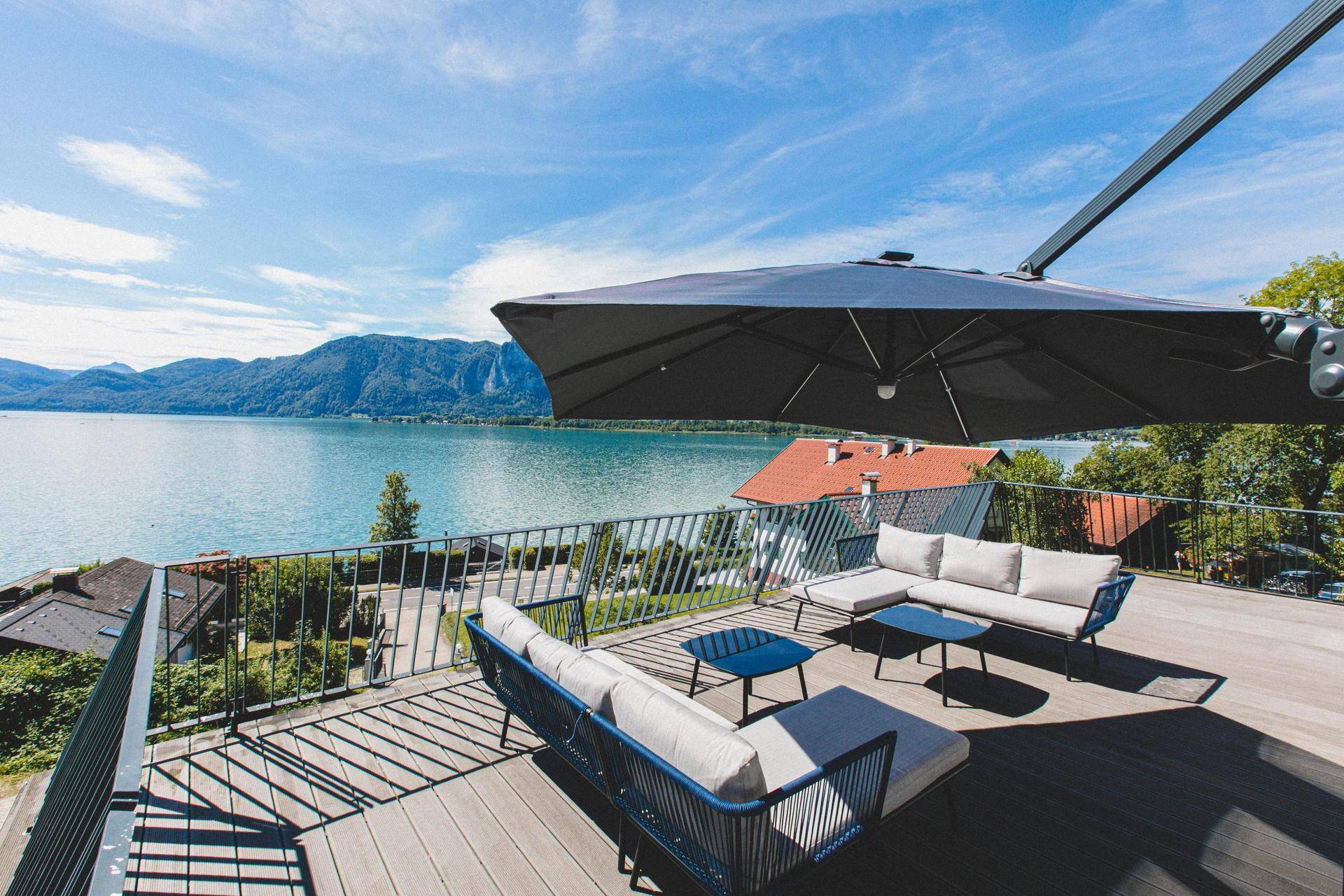 Geräumige Terrasse mit bequemen Lounge-Möbeln und großem Sonnenschirm, mit weitem Blick auf den See.