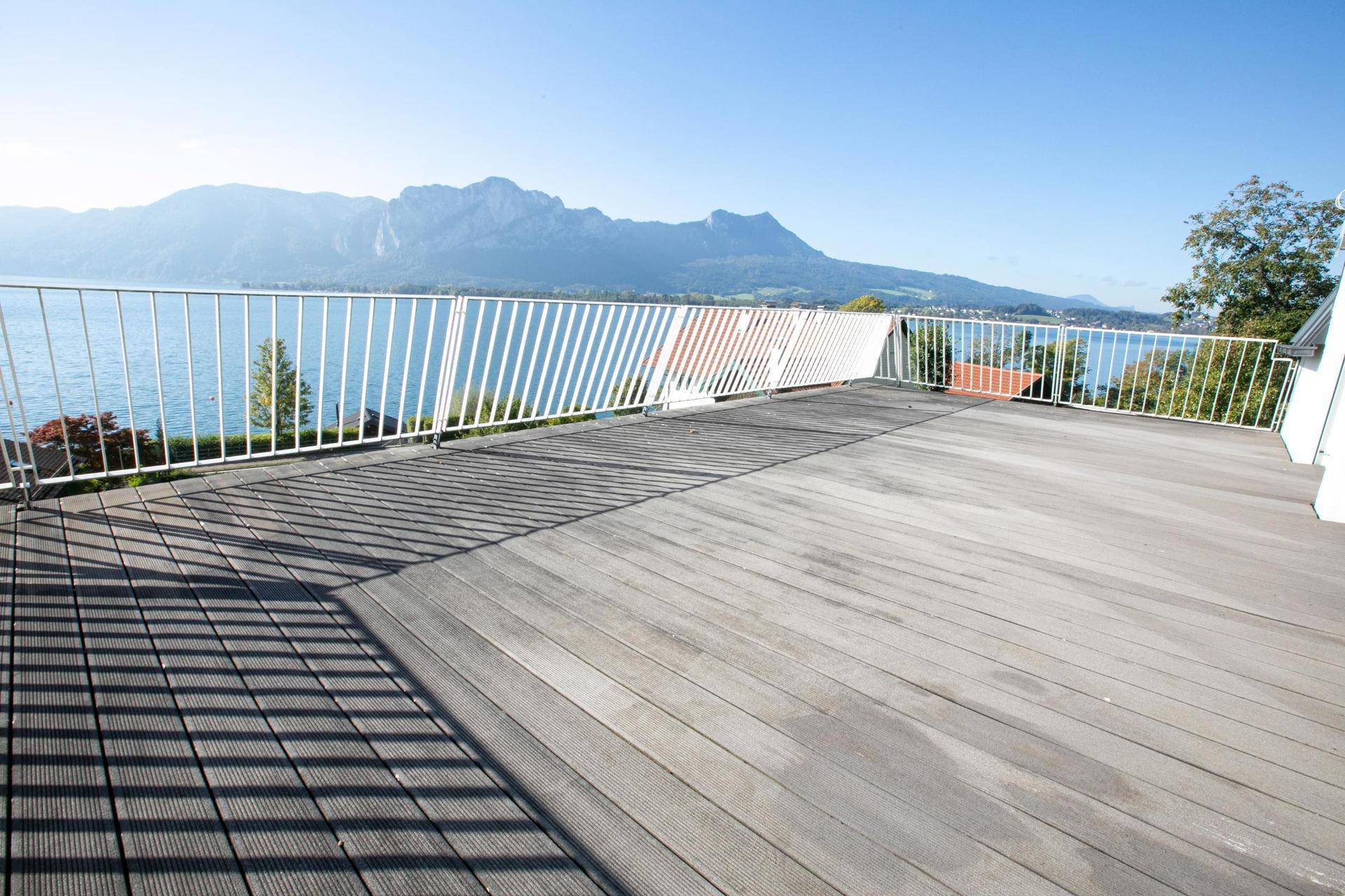 Weitläufige Holzterrasse mit weißem Geländer und Panoramablick auf den See und die umliegenden Berge.