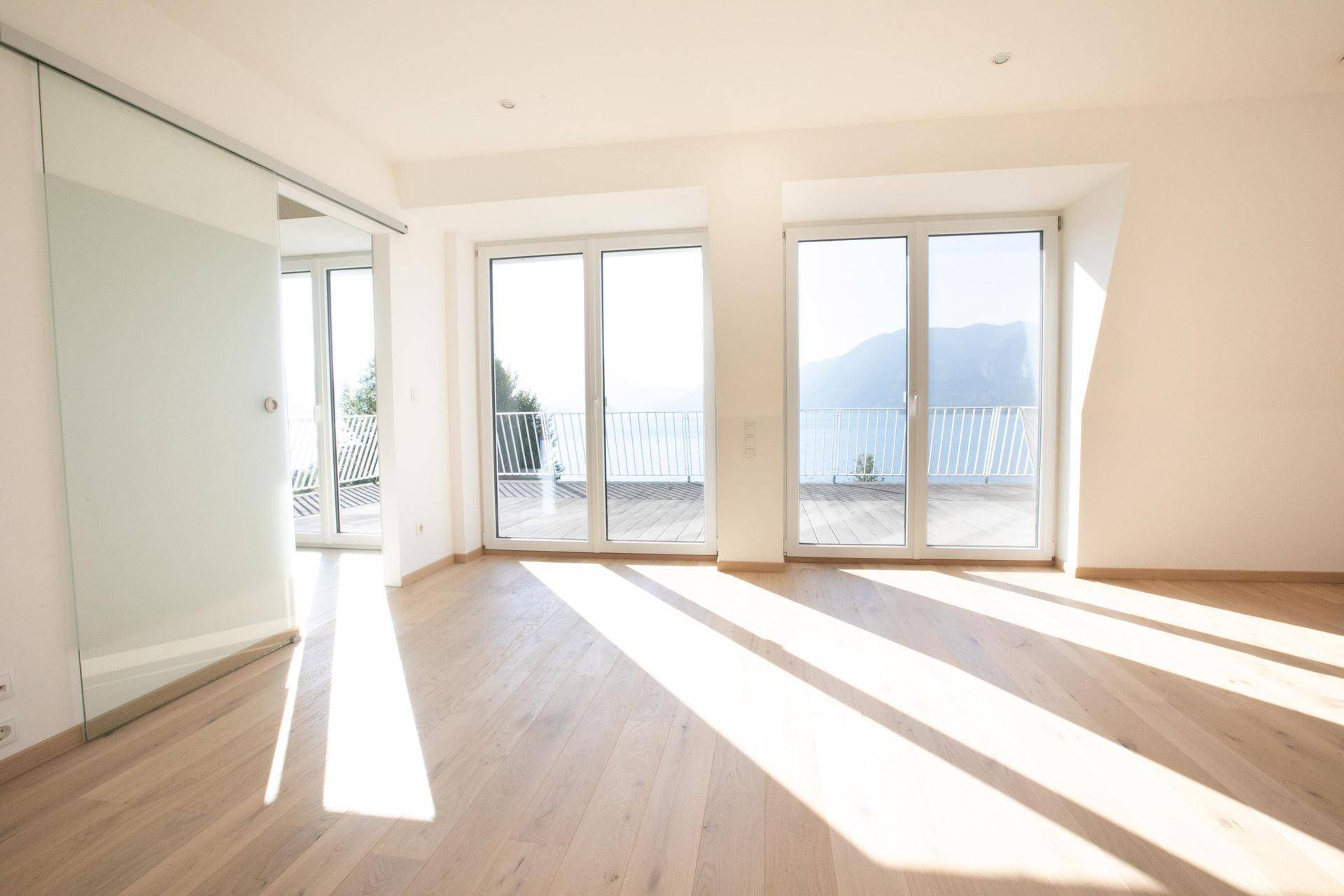 Lichtdurchflutetes Zimmer mit Holzboden und großen Fenstern, die Zugang zu einem Balkon mit Seeblick bieten.