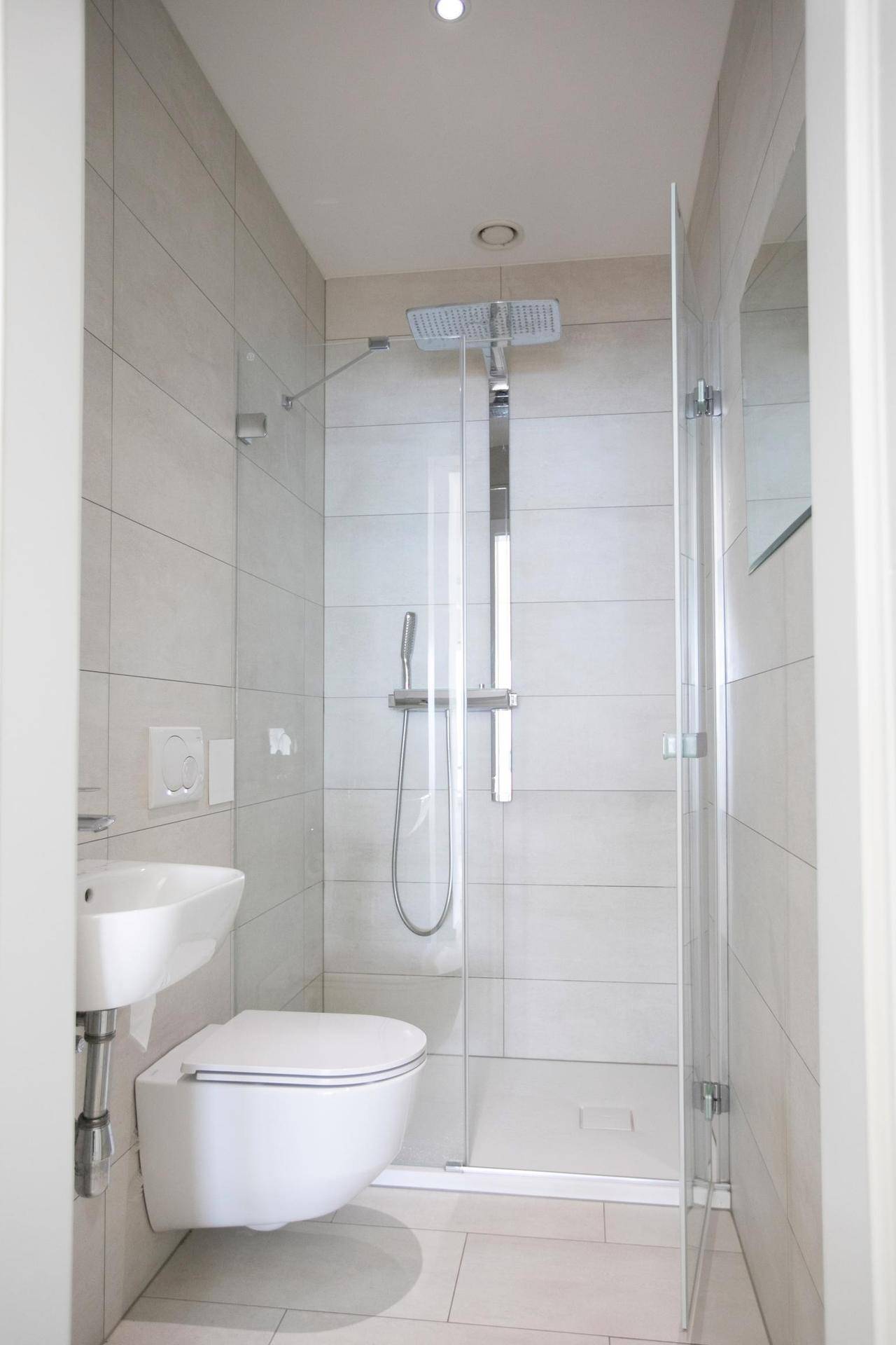 Kompaktes Badezimmer mit Dusche, wandhängendem WC und kleinem Waschbecken, hell gefliest.