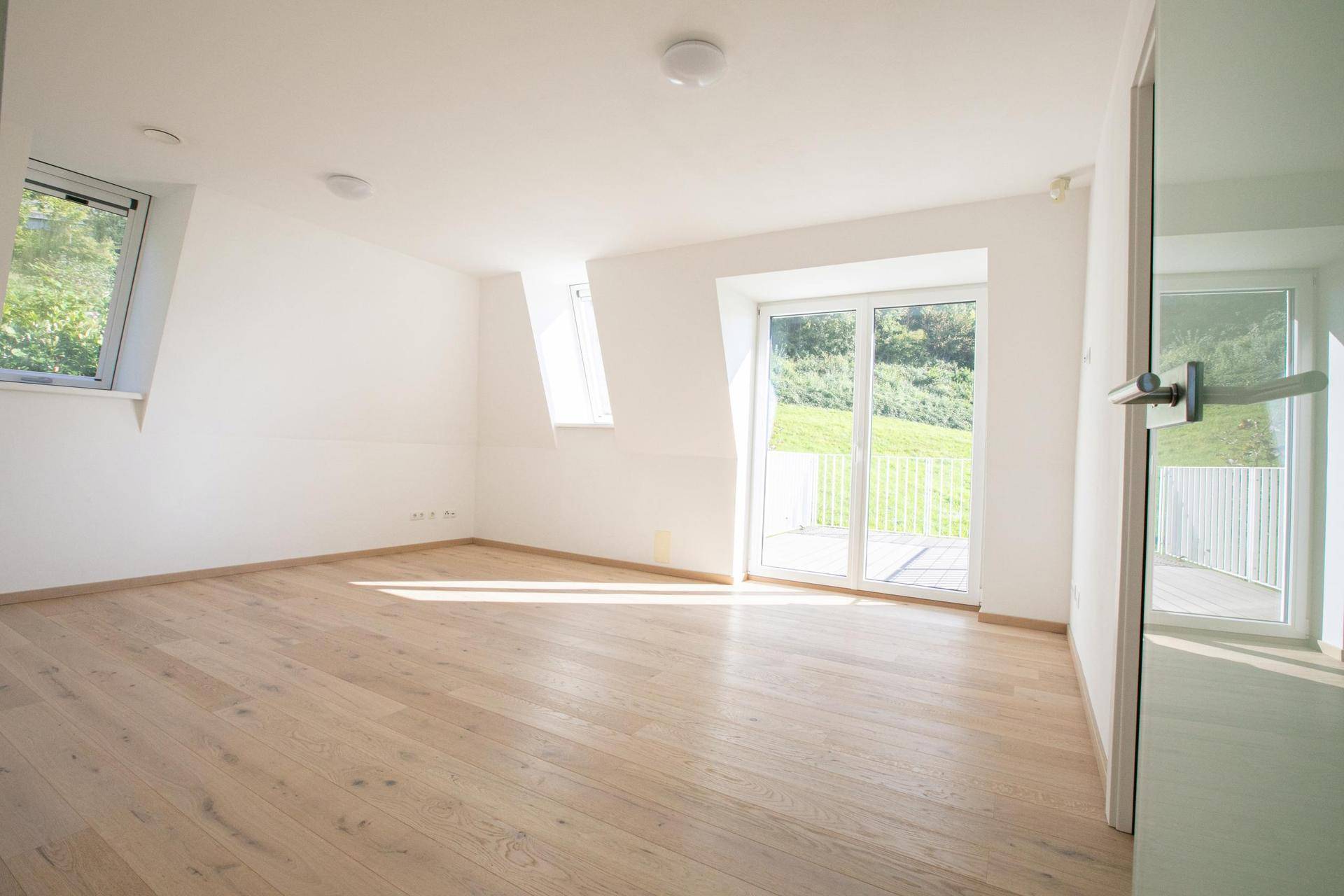 Helles Zimmer mit Holzboden, Dachfenstern und Balkonzugang, mit Blick auf die grüne Landschaft.