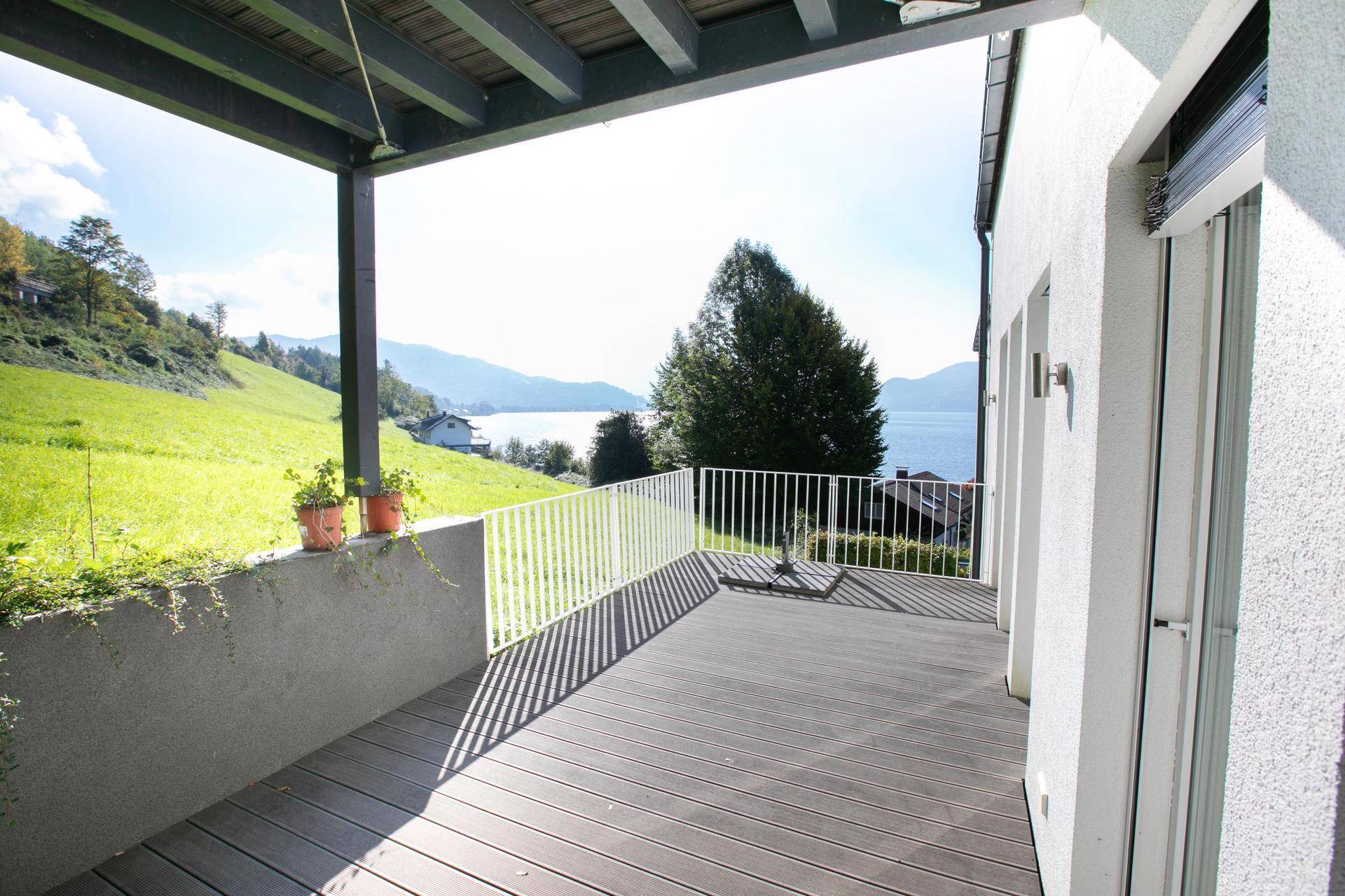 Überdachter Balkon mit weißem Geländer und sonnigem Ausblick auf die grünen Hügel und den See.