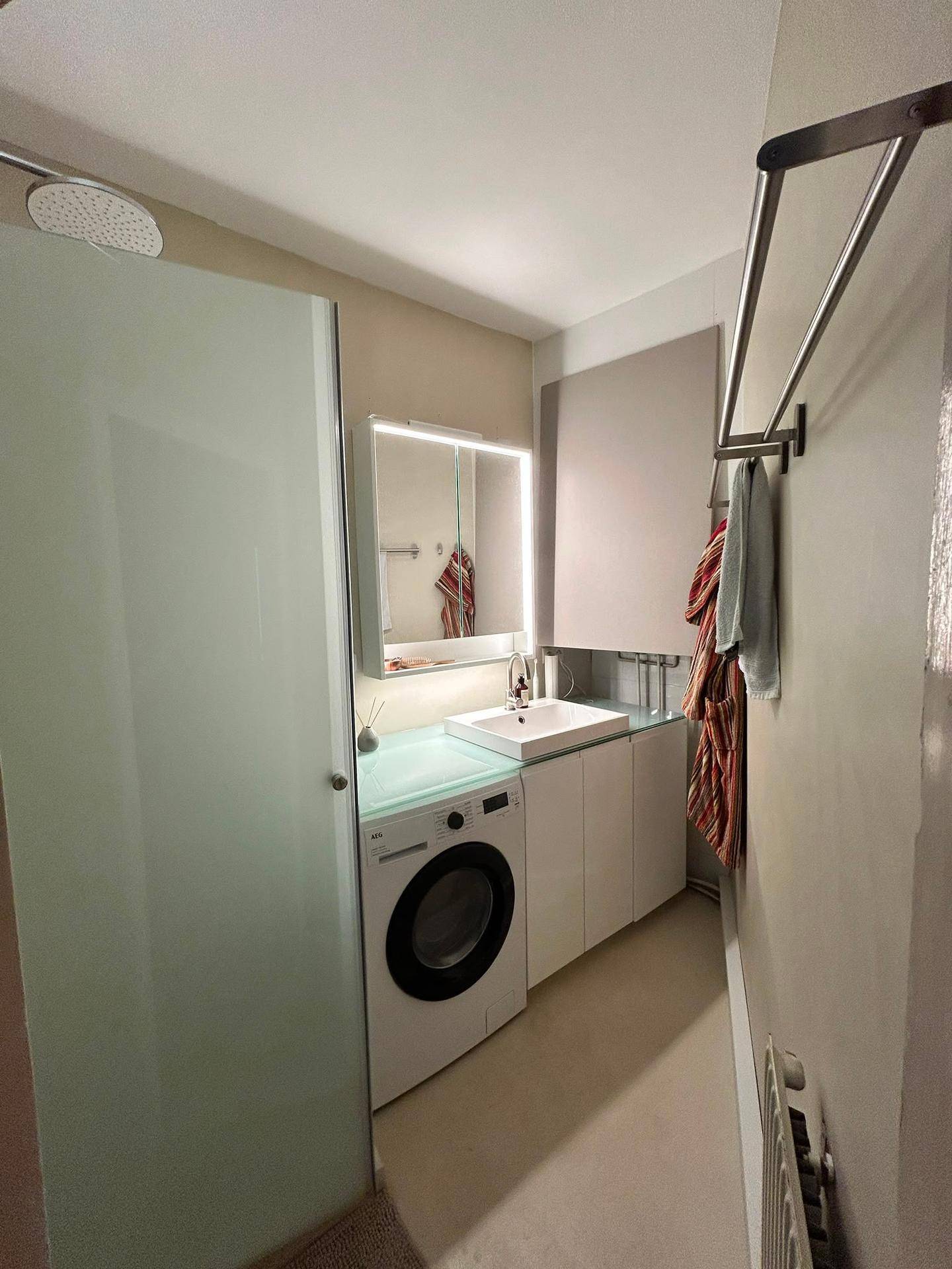 Modernes Badezimmer mit Dusche, Waschbecken, Waschmaschine und beleuchtetem Spiegelschrank.