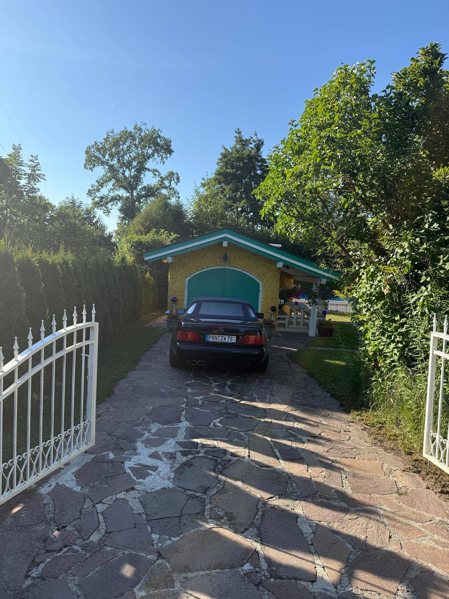 Einfahrt zum Grundstück mit weißem Tor, gepflastertem Weg und gelbem Haus im Hintergrund.