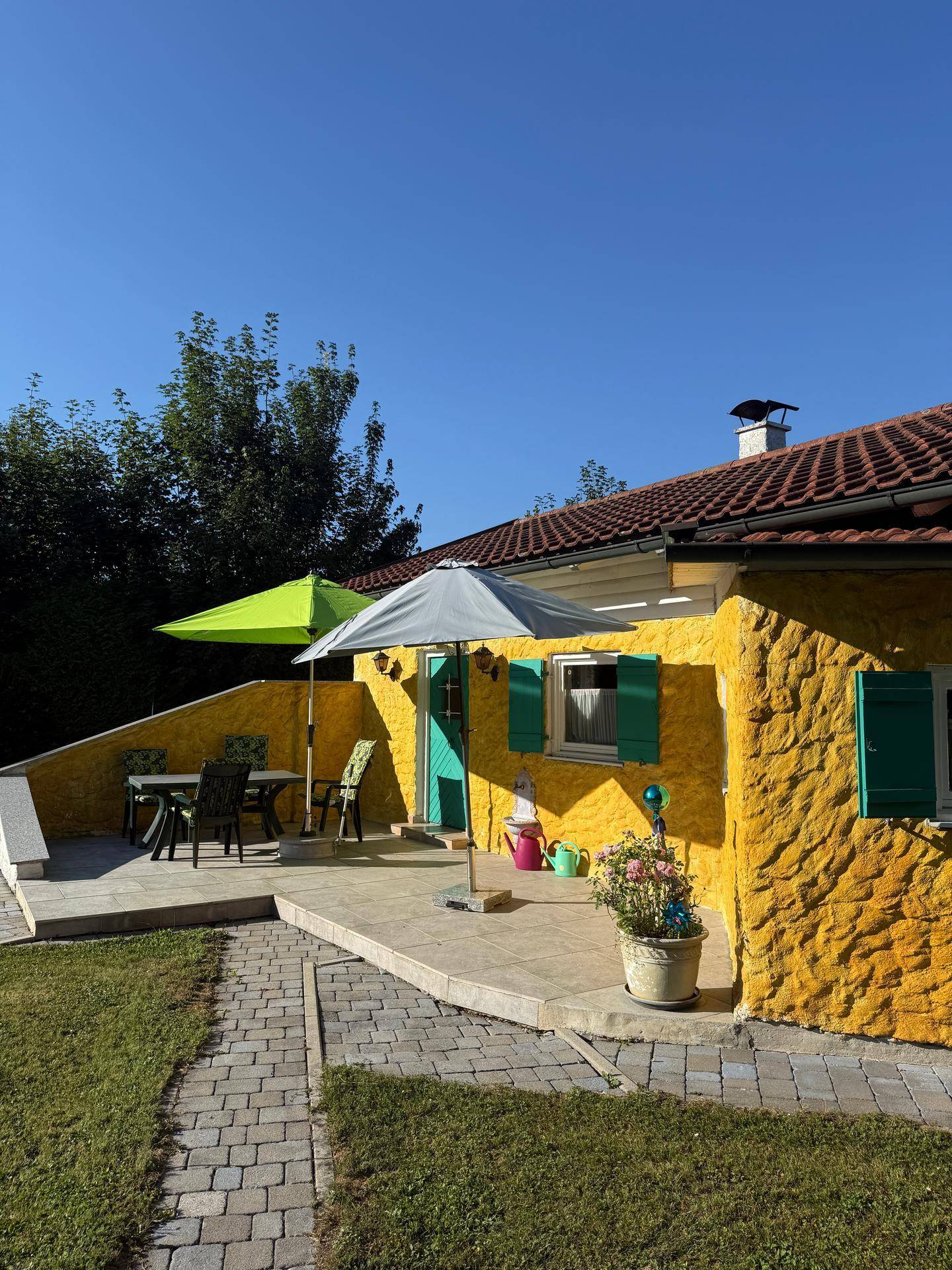 Sonniger Terrassenbereich am gelben Haus mit grünen Fensterläden und Gartenmöbeln.