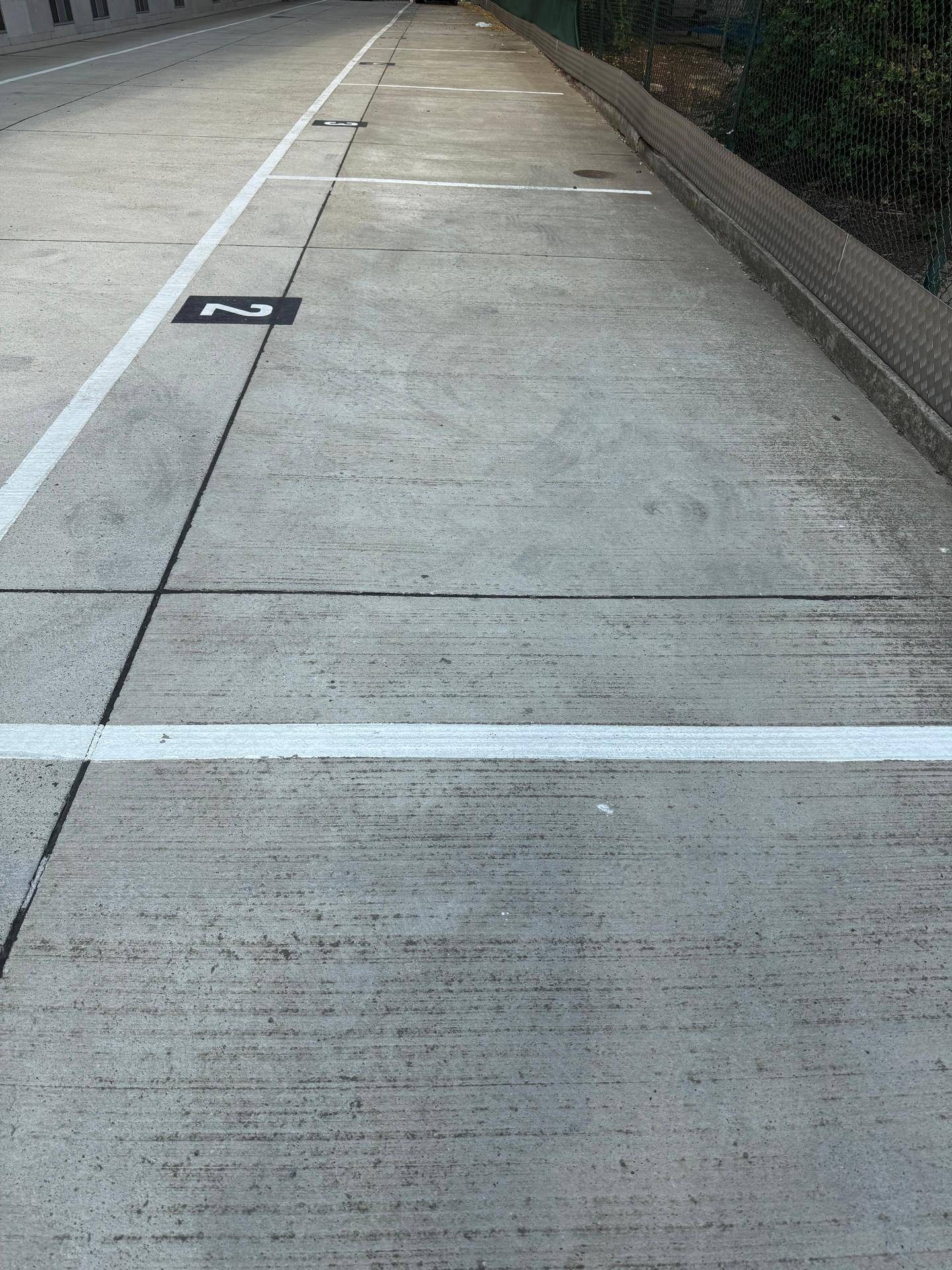 Langer Außenparkplatz mit mehreren markierten Stellplätzen und einem Zaun im Hintergrund.