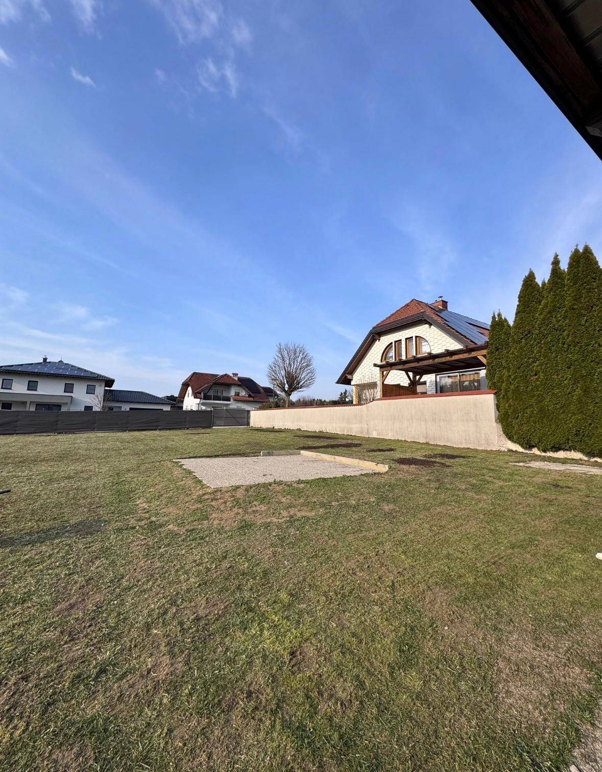 Weitläufige Rasenfläche mit einem Haus im Hintergrund, das eine Holzpergola und Solarpaneele aufweist.