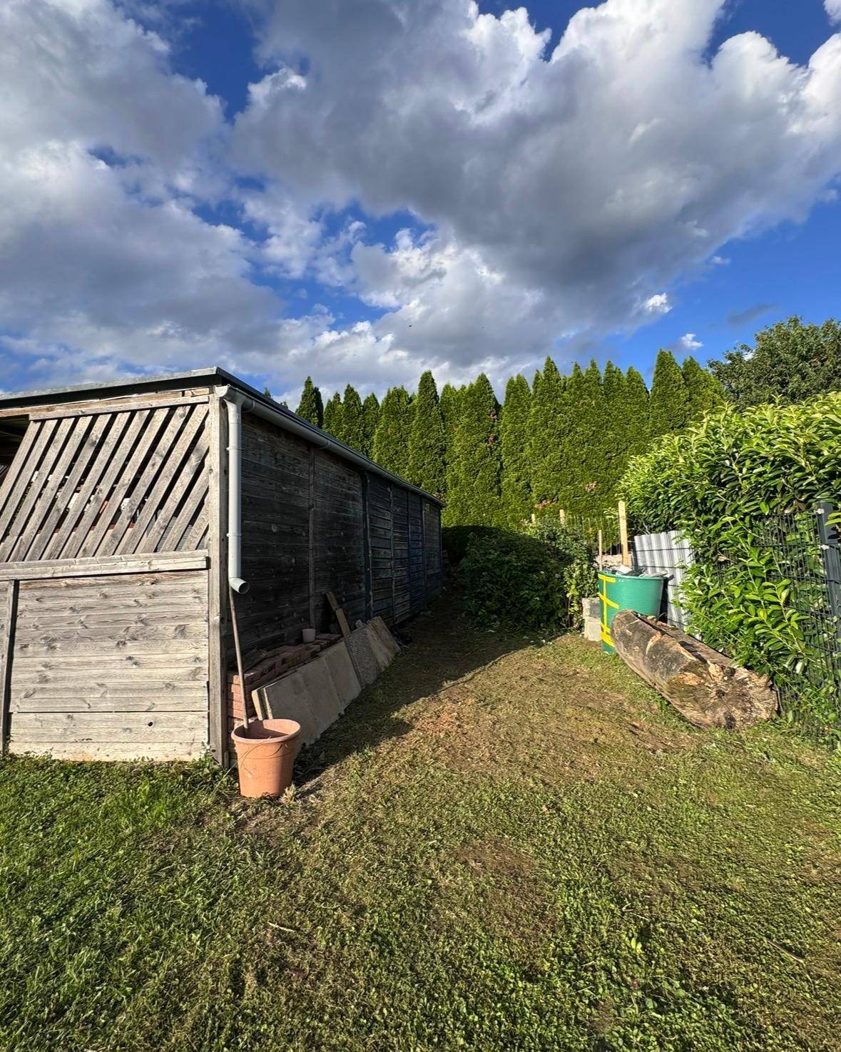 Rustikales Holzgartenhaus neben einer dichten Hecke und Abfallbehältern unter bewölktem Himmel.