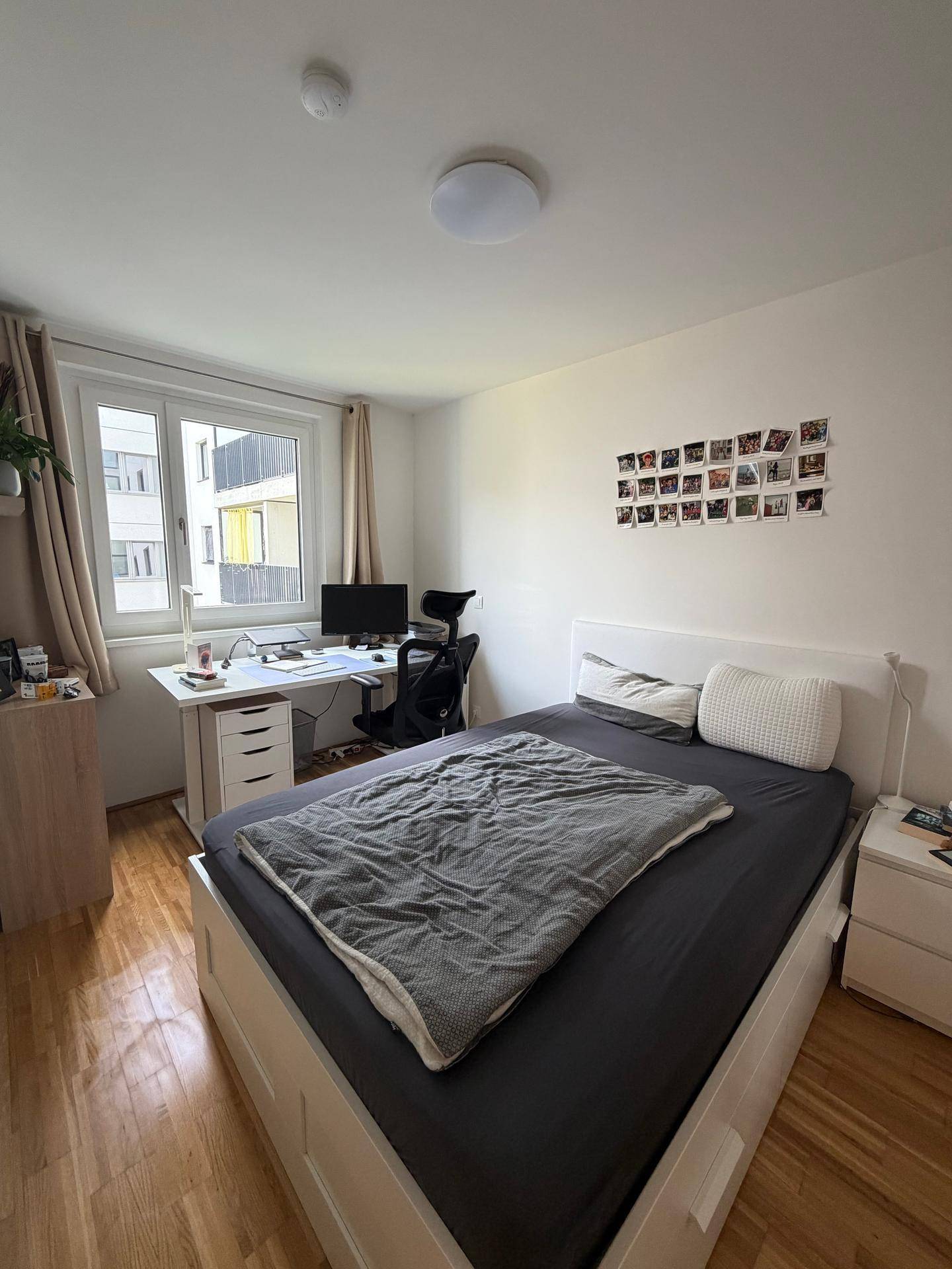Helles Schlafzimmer mit Schreibtisch am Fenster, weißem Bett und Holzboden.