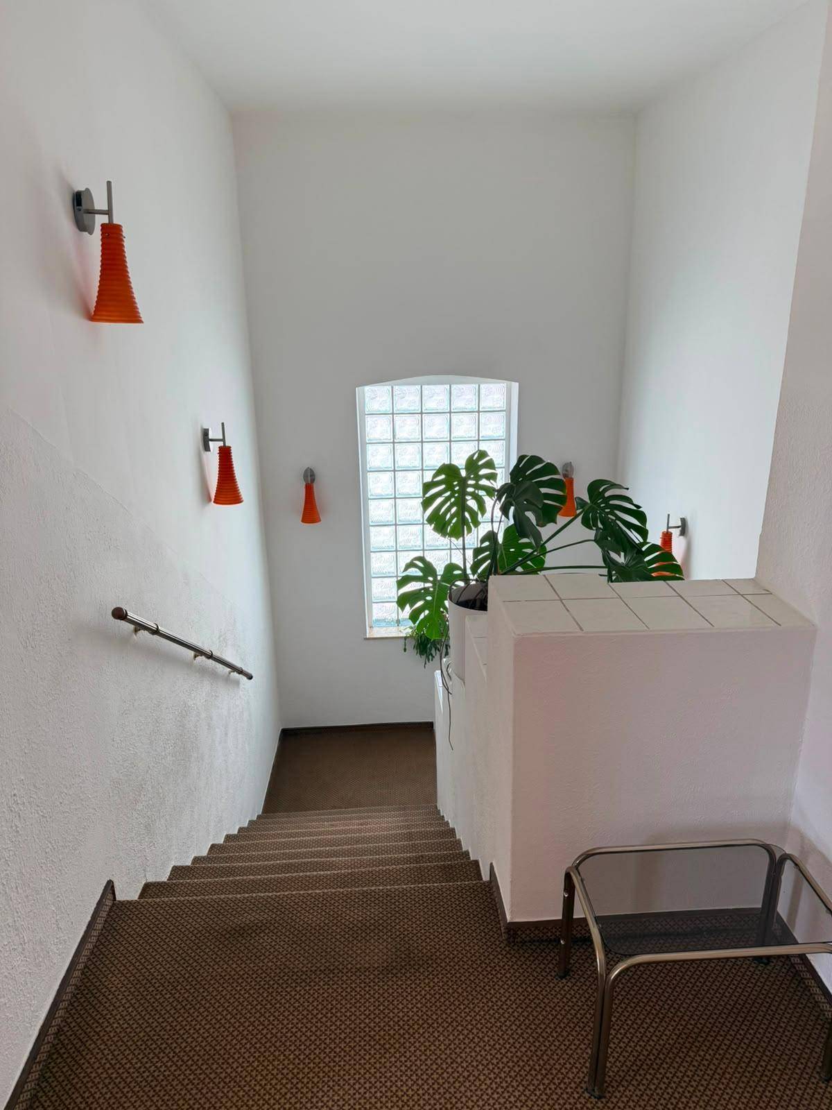 Helles Treppenhaus mit Glasbausteinfenster, orangefarbenen Wandleuchten und einer großen Pflanze.