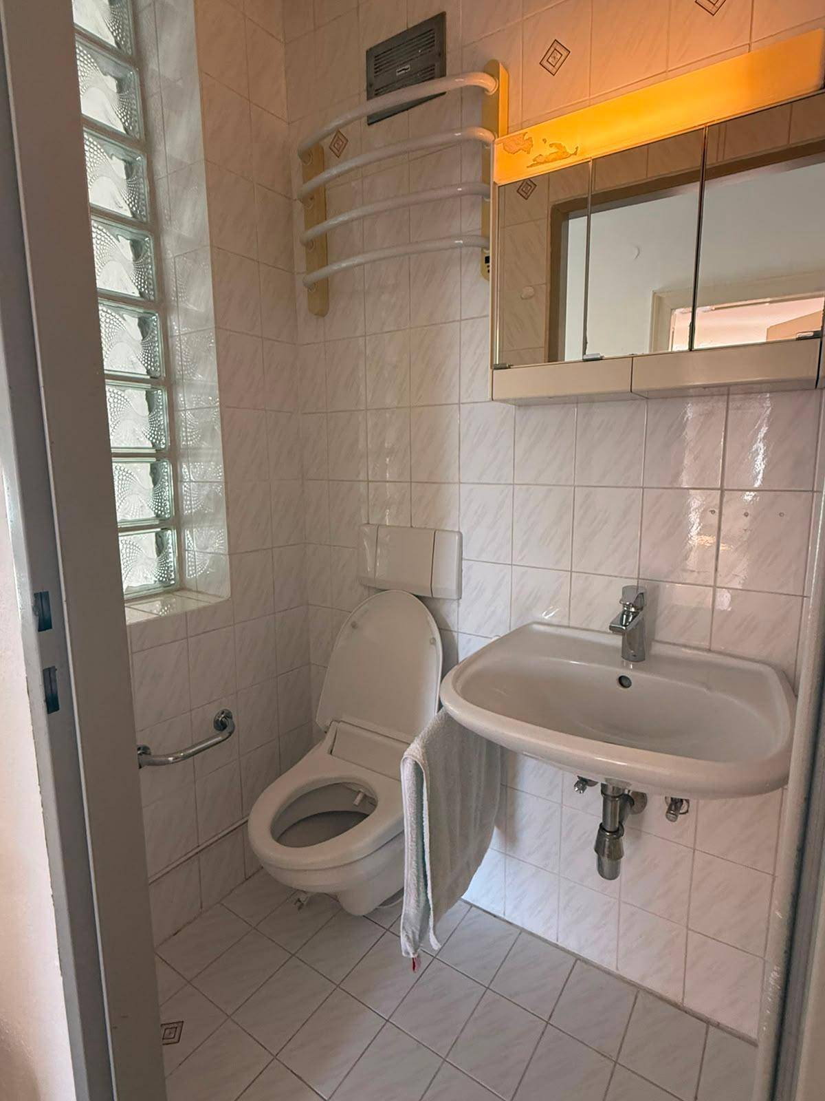 Badezimmer mit Toilette, Waschbecken, Spiegelschrank und einem Glasbausteinfenster.