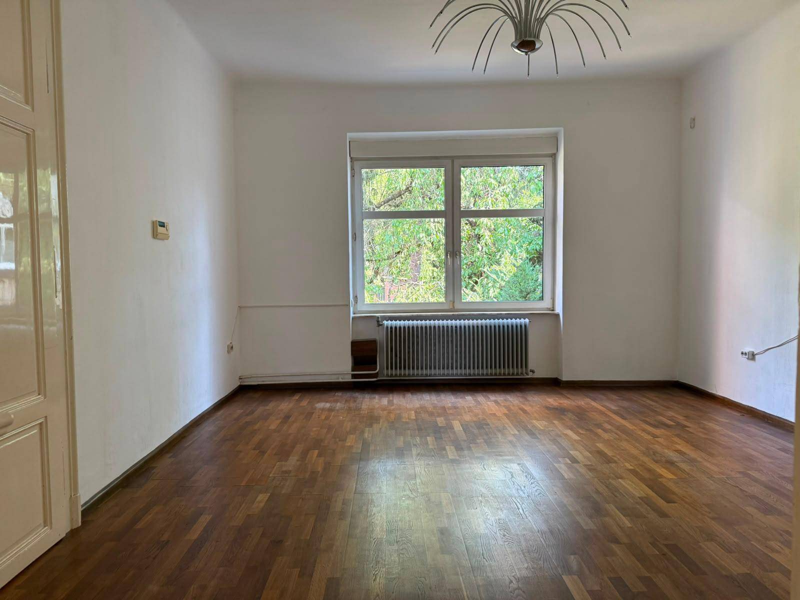 Helles, leeres Zimmer mit Parkettboden, weißen Wänden und großem Fenster mit Blick ins Grüne.