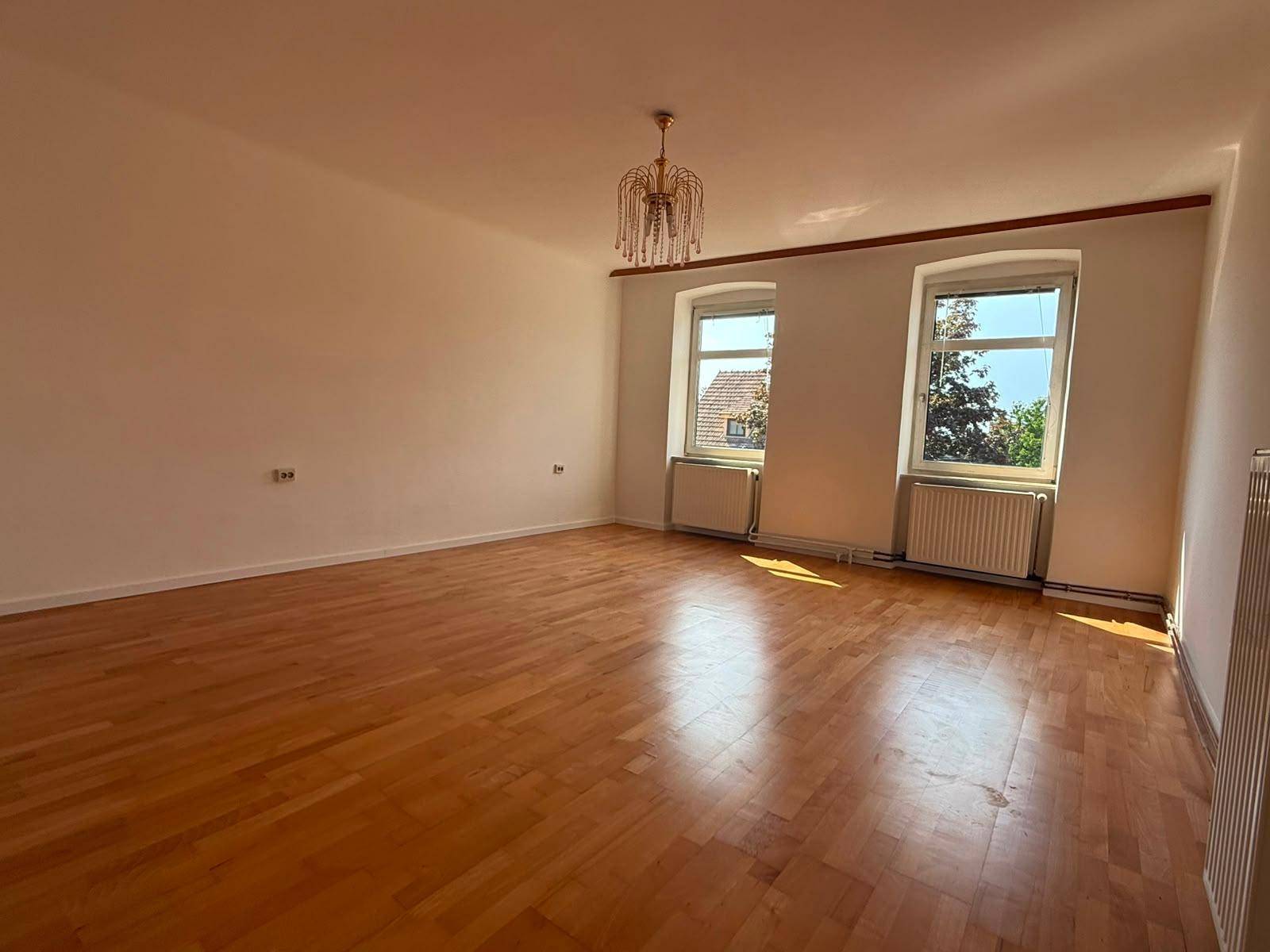 Geräumiges, leeres Wohnzimmer mit hellem Parkettboden, weißen Wänden und einem Kronleuchter.