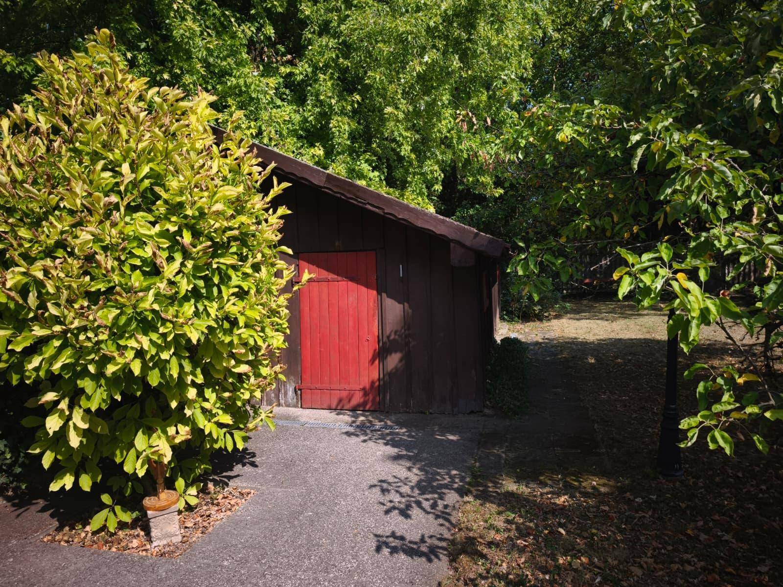 Kleines, rustikales Gartenhaus aus Holz mit roter Tür, umgeben von dichtem Grün und Bäumen.