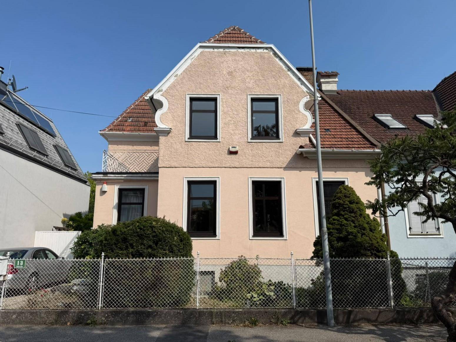 Ansicht des Hauses von der Straße mit hellrosa Fassade, dunklen Fenstern und einem kleinen Vorgarten.