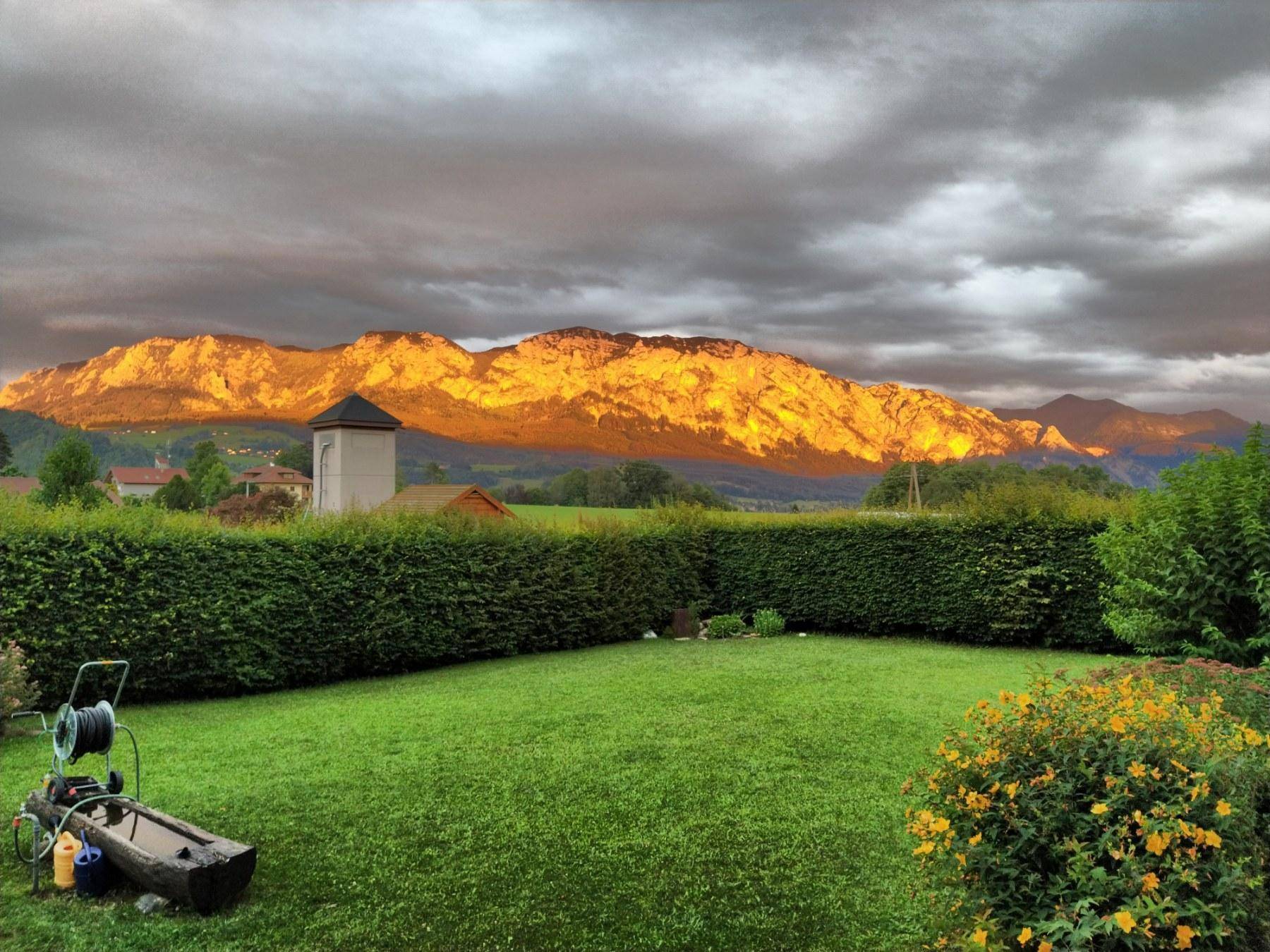 Gepflegter Garten mit Rasenfläche und Hecke, im Hintergrund die von der Sonne beleuchteten Berge.