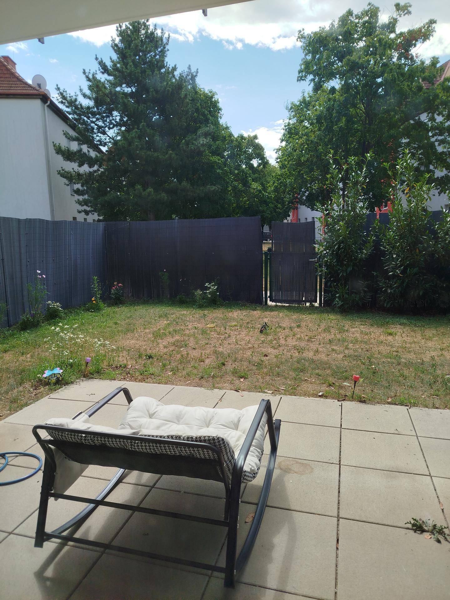 Grüner Gartenbereich mit Rasenfläche, Zaun und einer gepflasterten Terrasse mit Sitzgelegenheit.