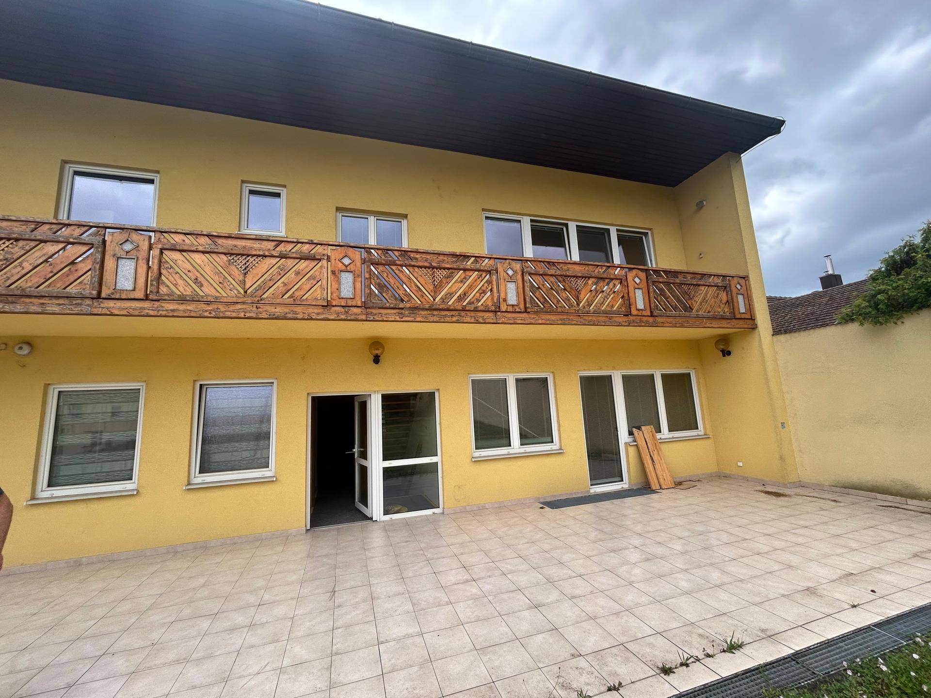 Hinteransicht des Gebäudes mit großem Holzbalkon und einer gefliesten Terrasse im Erdgeschoss.
