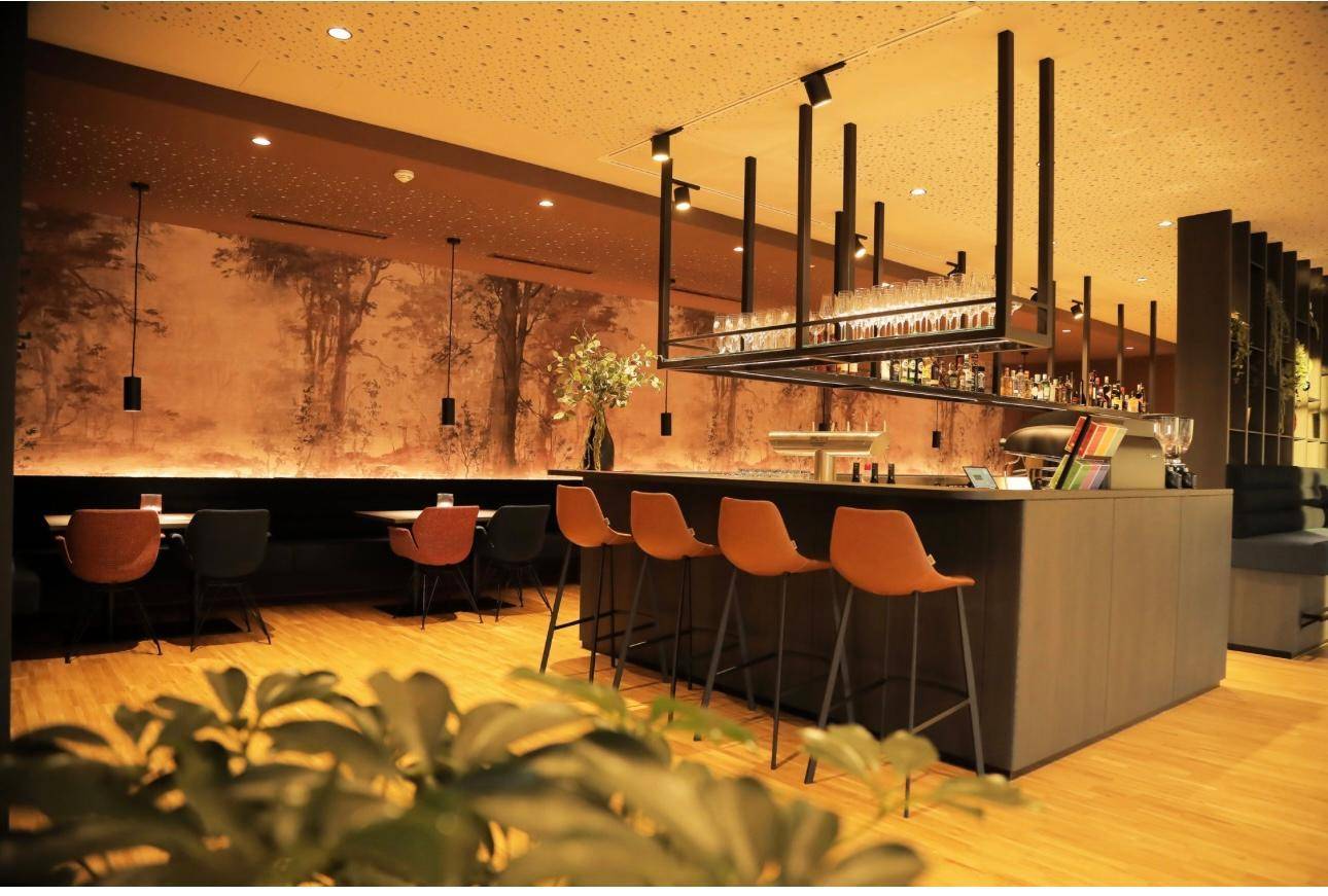 Gesamtansicht des modernen Restaurant- und Barbereichs mit dunkler Theke, Barhockern und einer Waldtapete.