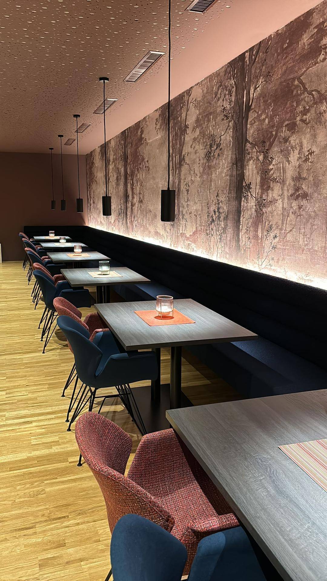 Langer Restaurantbereich mit Reihen von Tischen und Stühlen, gepolsterten Bänken und einer Waldtapete.
