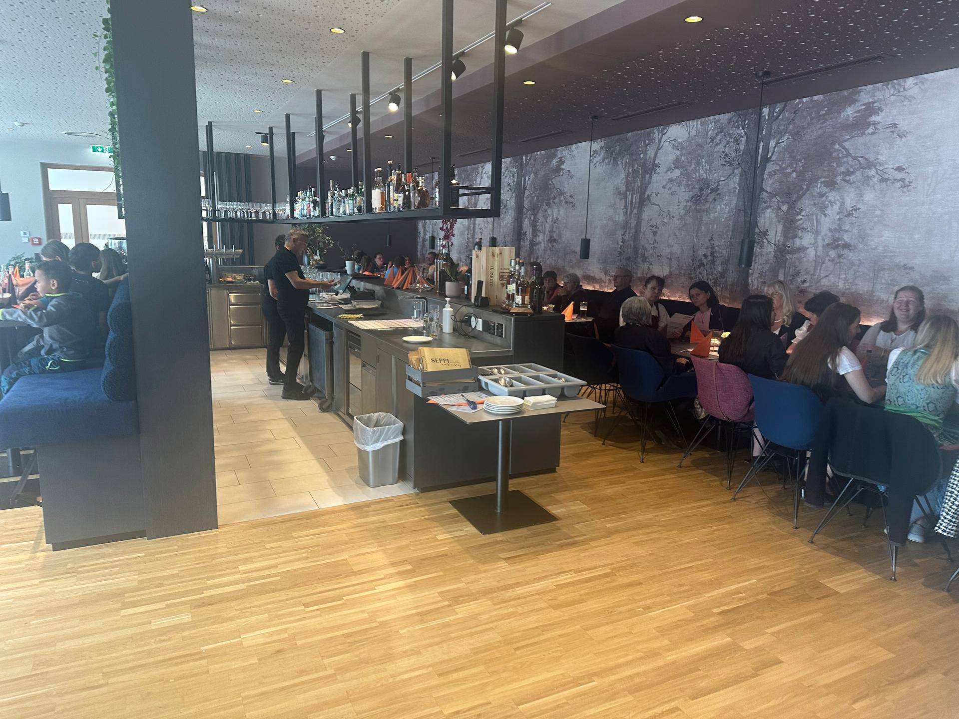 Belebtes Restaurant mit Gästen, einer offenen Servicetheke und Holzboden, akzentuiert durch eine Waldtapete.