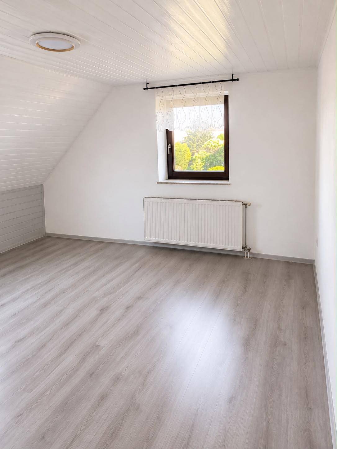 Helles Zimmer mit weißer Holzdecke, Laminatboden und Fenster mit Blick ins Grüne.