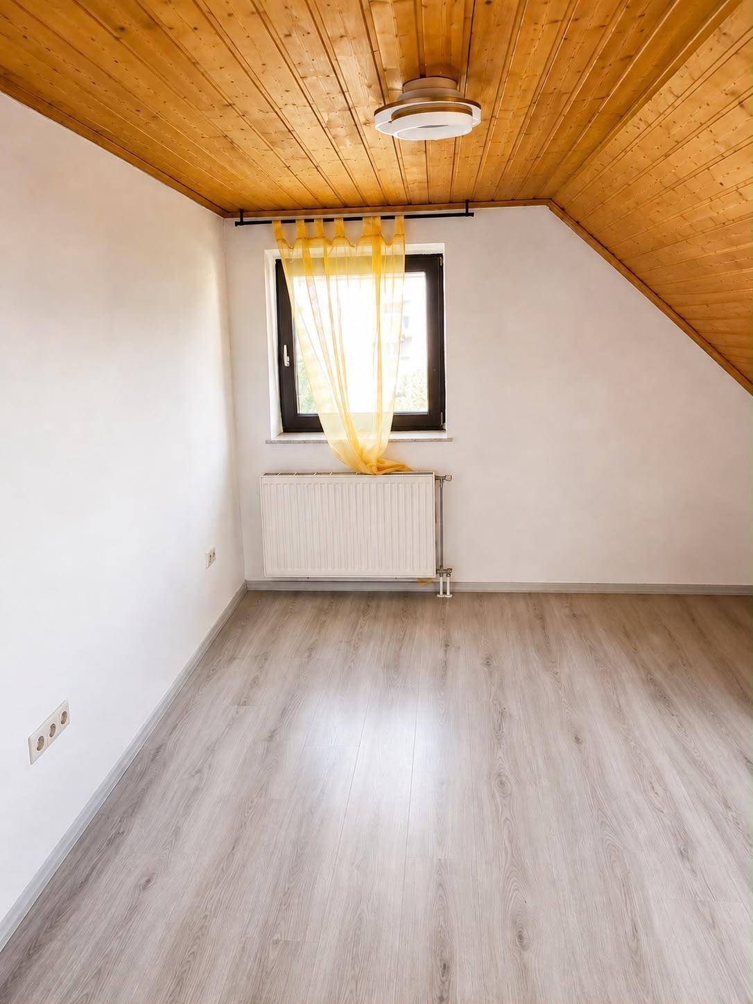 Leeres Zimmer mit Holzdecke, Laminatboden und Fenster mit gelbem Vorhang.