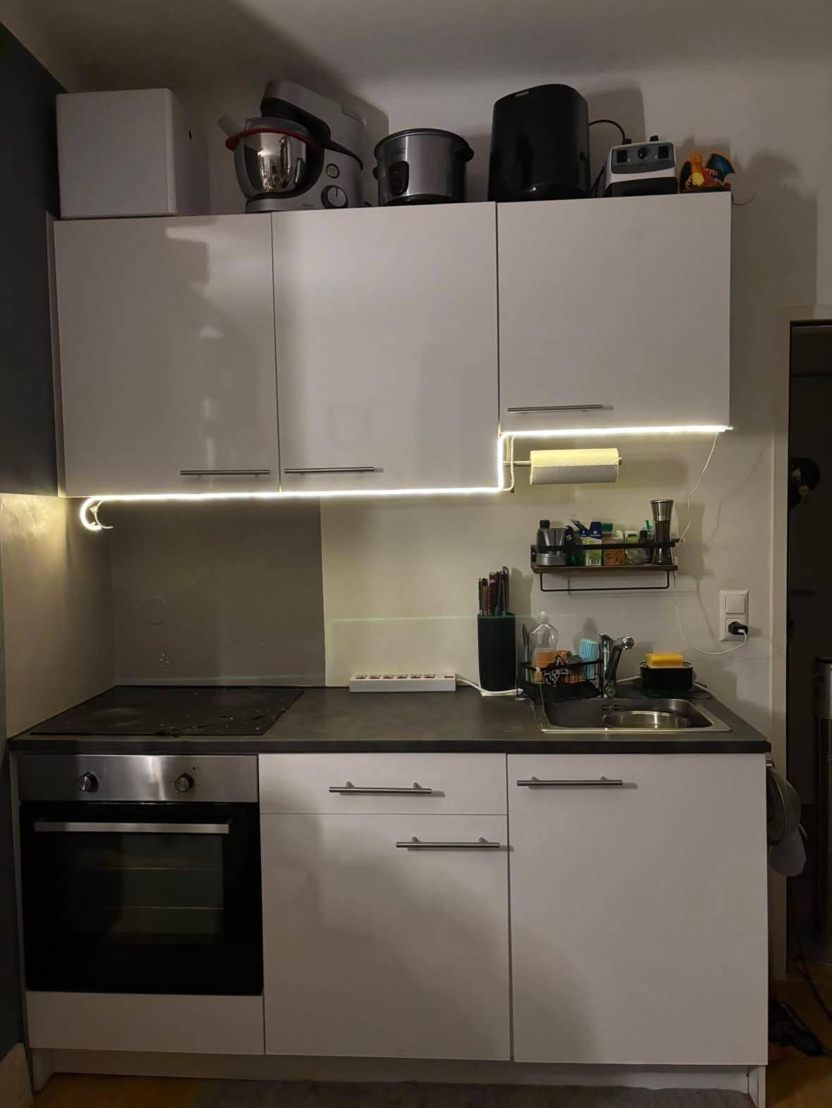 Kleine, funktionale Küchenzeile mit weißen Schränken, Kochfeld, Backofen und Spüle.