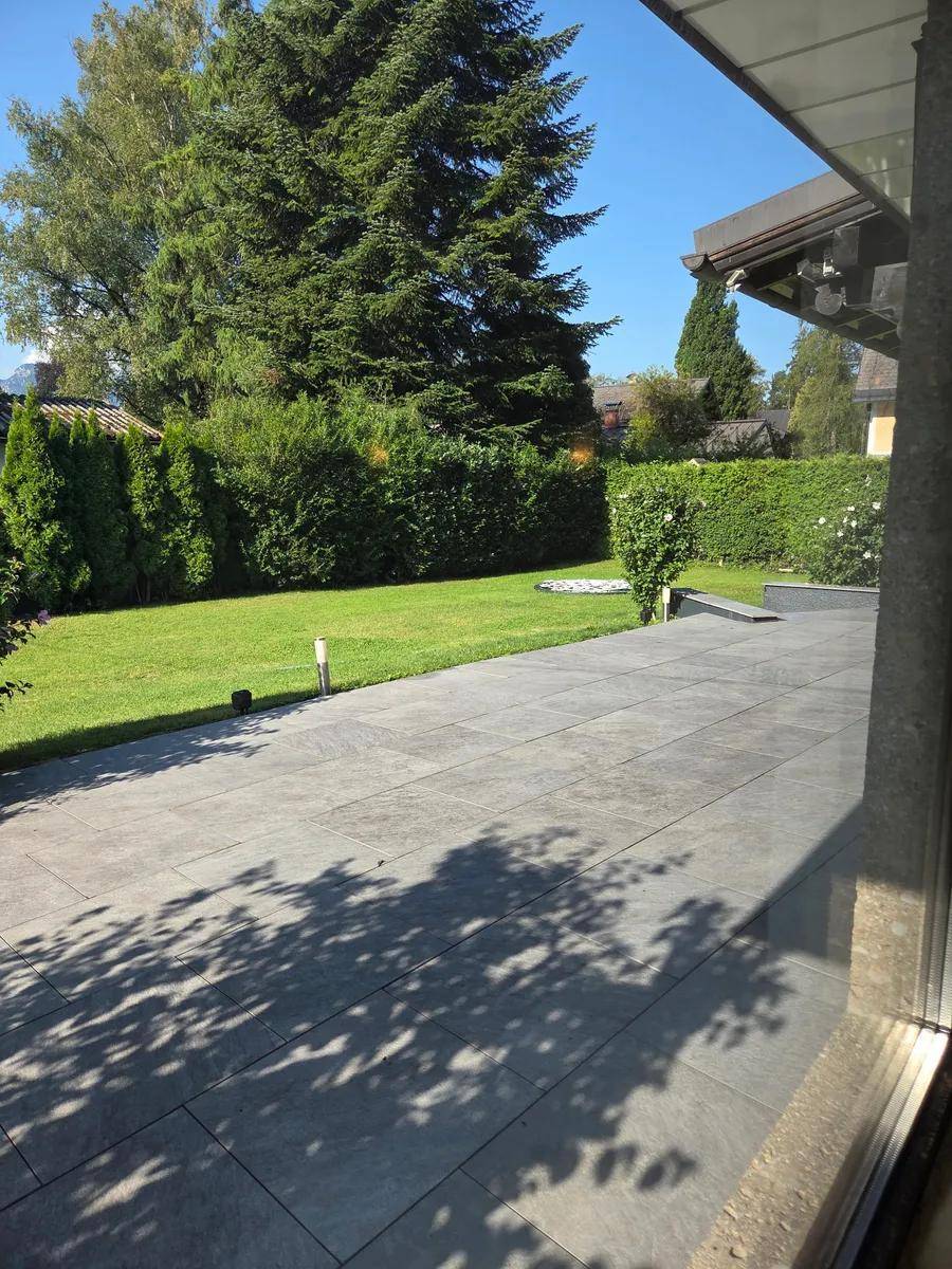 Große geflieste Terrasse mit Rasenfläche, Hecken und Bäumen im Hintergrund.