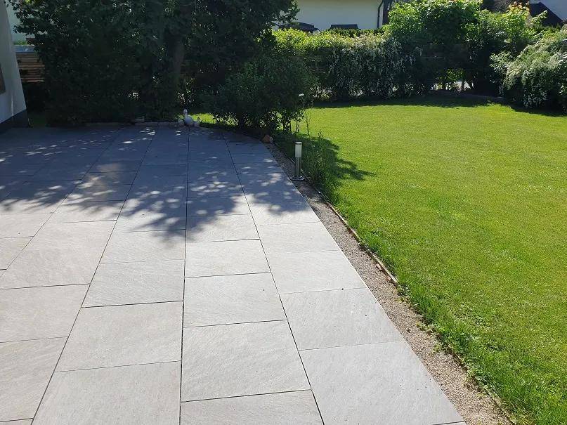 Gepflegte Terrasse mit grauen Fliesen, angrenzendem Rasen und grünen Hecken.