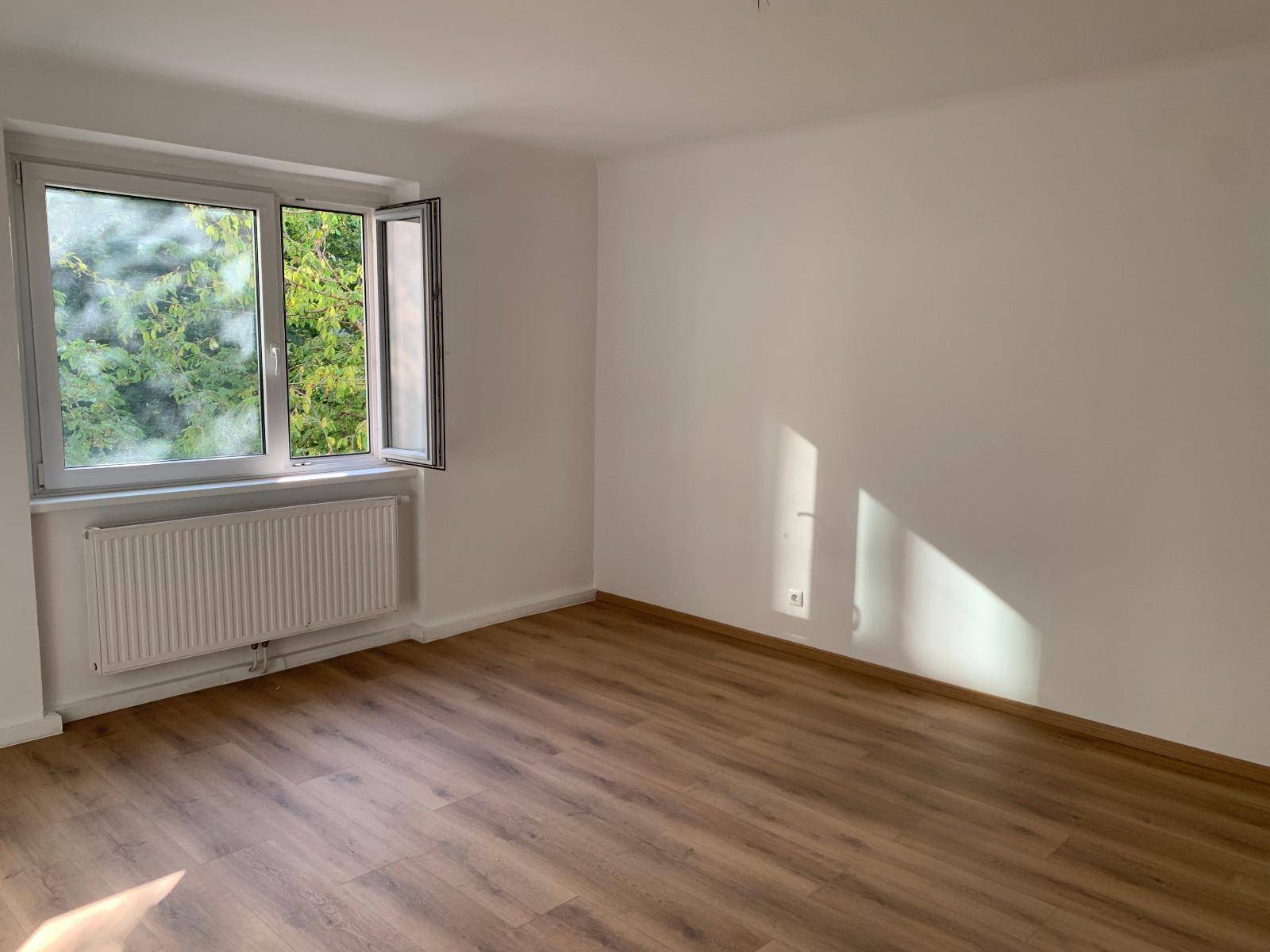 Helles Zimmer mit Laminatboden und geöffnetem Fenster, das einen Blick ins Grüne bietet.