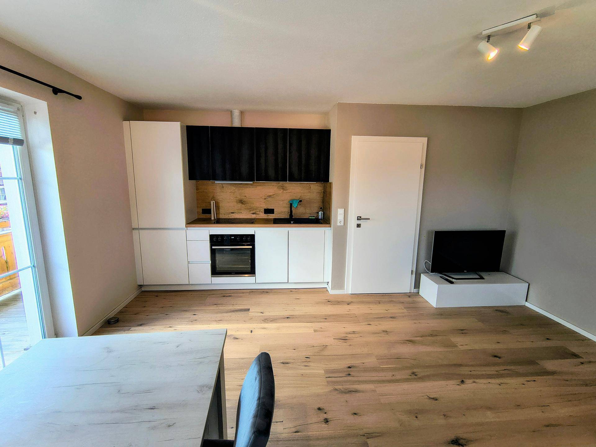 Heller offener Wohn- und Essbereich mit moderner Einbauküche, Holzboden und Zugang zum Balkon.