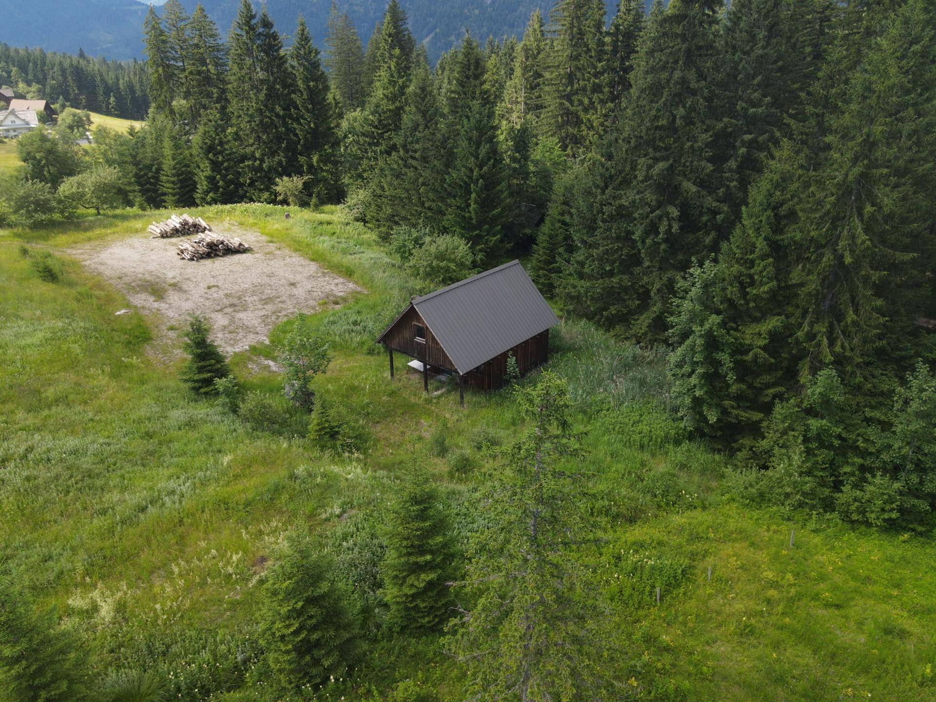 Luftaufnahme einer abgelegenen Holzhütte in einer Waldlichtung mit Holzstapeln auf dem Grundstück.
