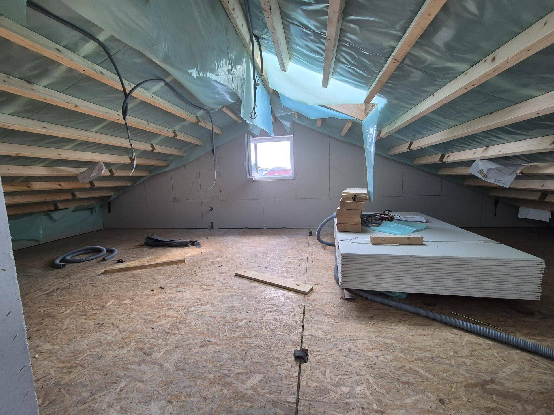 Dachgeschoss im Rohbau mit sichtbaren Holzbalken, Isolierung und einem kleinen Fenster.