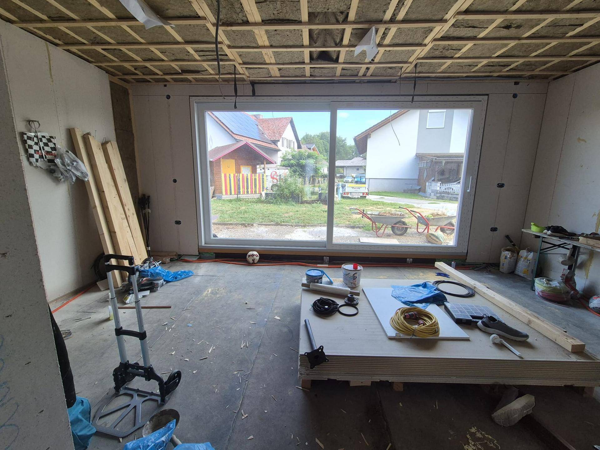 Innenraum im Rohbau mit großer Fensterfront und Blick auf den Garten, Decke mit Isolierung.
