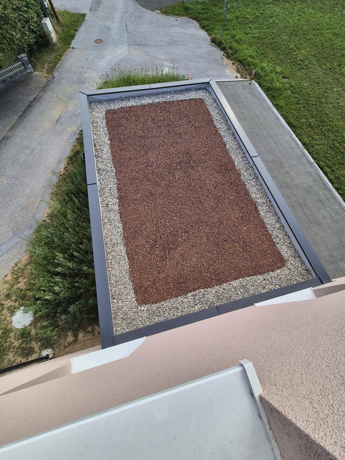 Draufsicht auf ein Flachdach mit Kies und einer begrünten Fläche, angrenzend an eine Straße.
