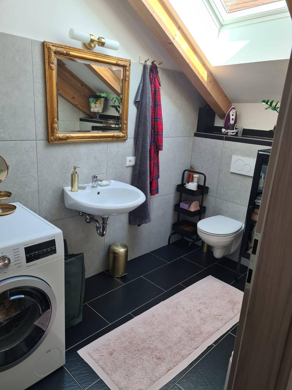 Modernes Badezimmer im Dachgeschoss mit grauen Fliesen, Waschmaschine und Tageslicht durch das Dachfenster.