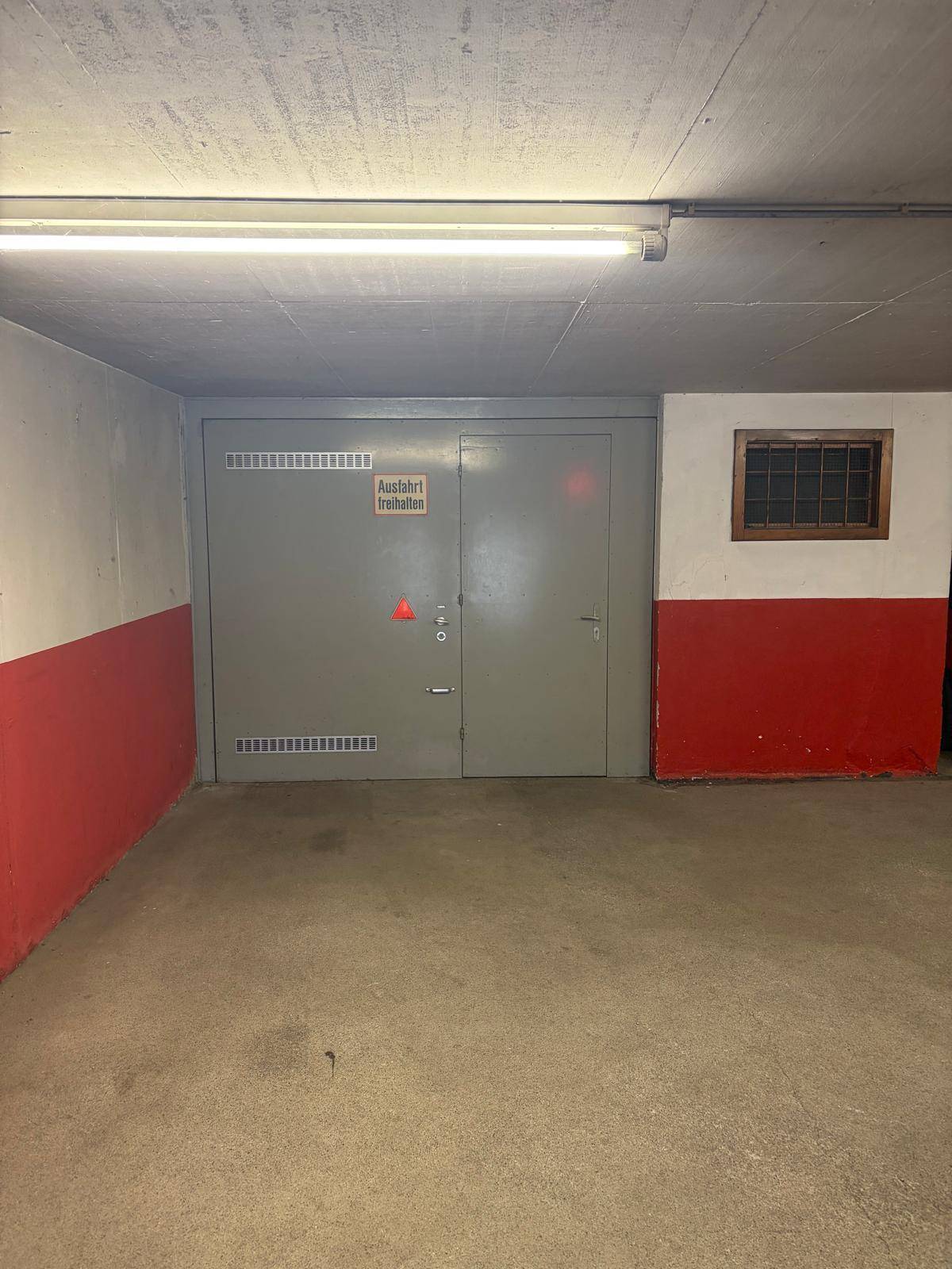 Tiefgarage oder Kellerraum mit Betonboden, grauen Türen und rot-weiß gestrichenen Wänden.
