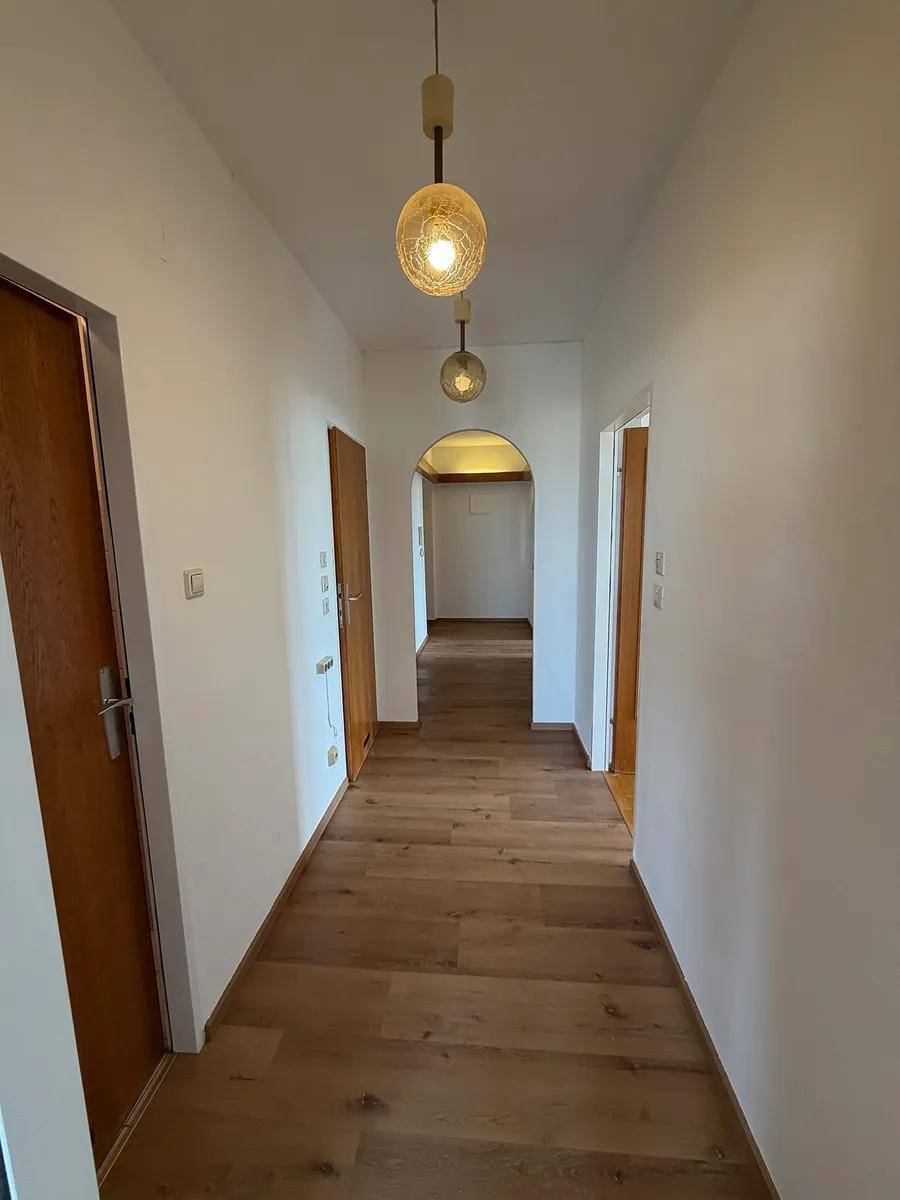 Langer Flur mit Holzboden, weißen Wänden und sichtbaren Holzbalken, beleuchtet von zwei Pendelleuchten.